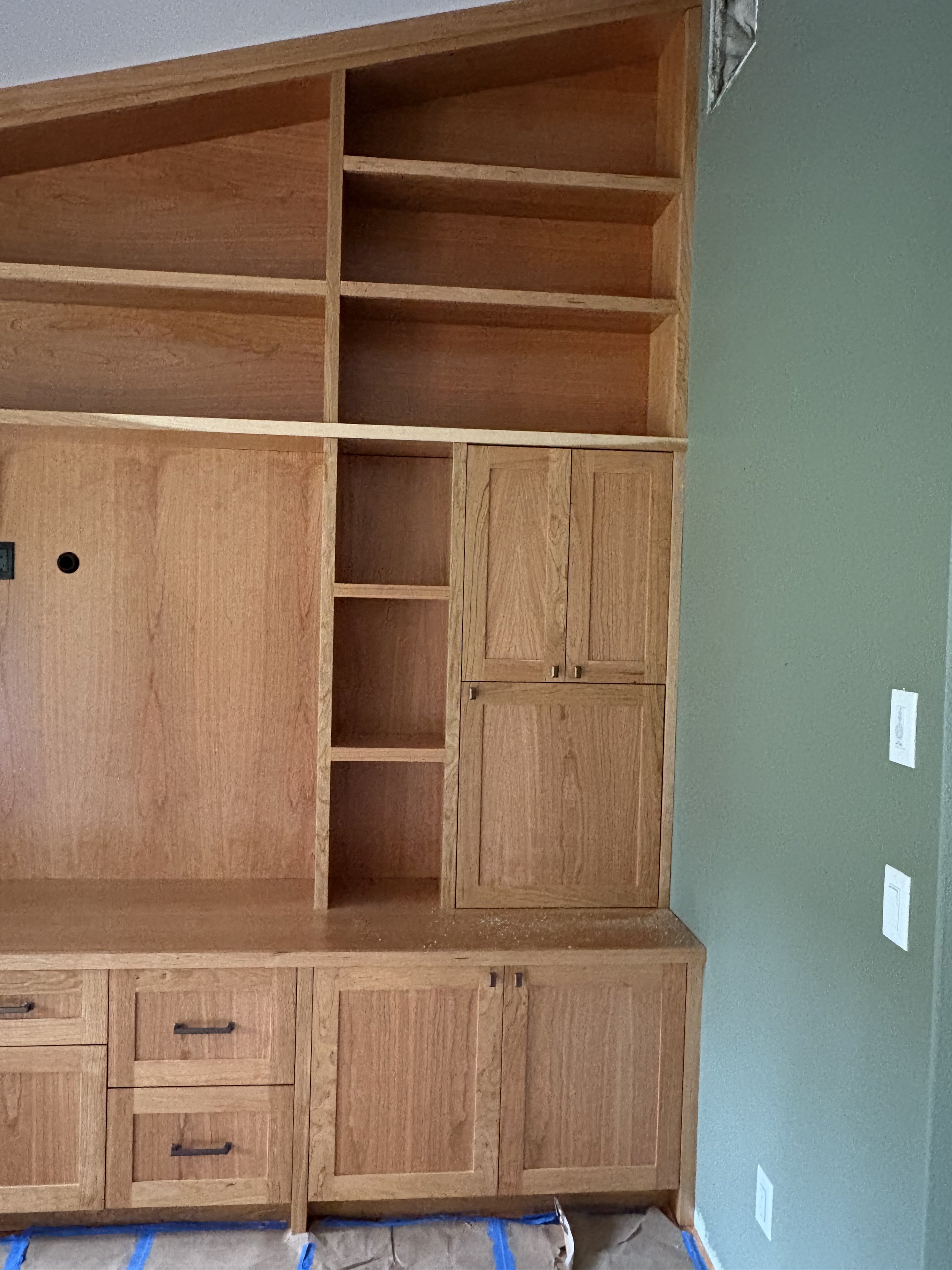 A wooden built-in cabinet with open shelves, drawers, and closed cabinet doors, installed against a green wall in a room under construction.