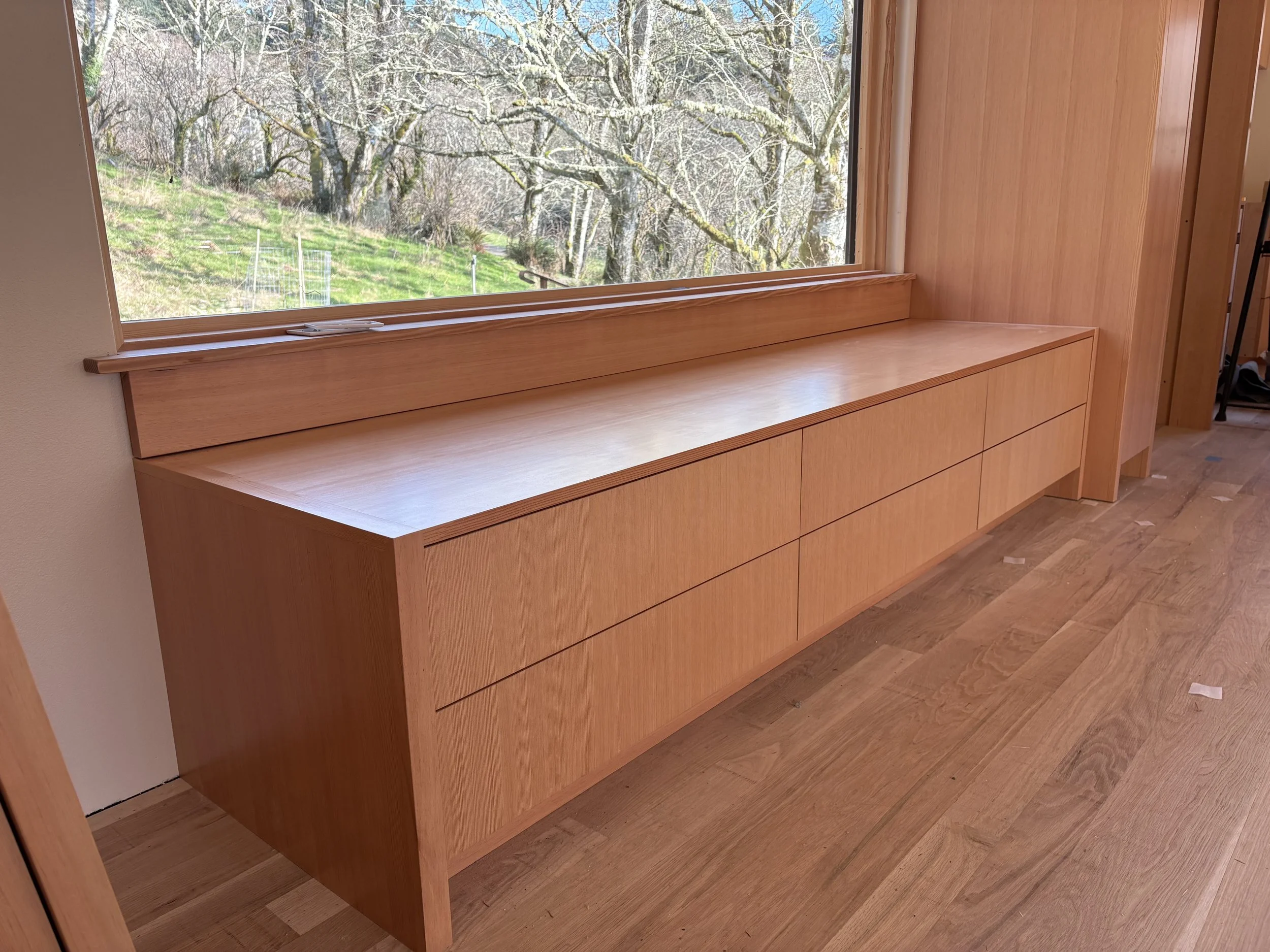 Wooden built-in cabinet with drawers beneath a large window overlooking trees outside.