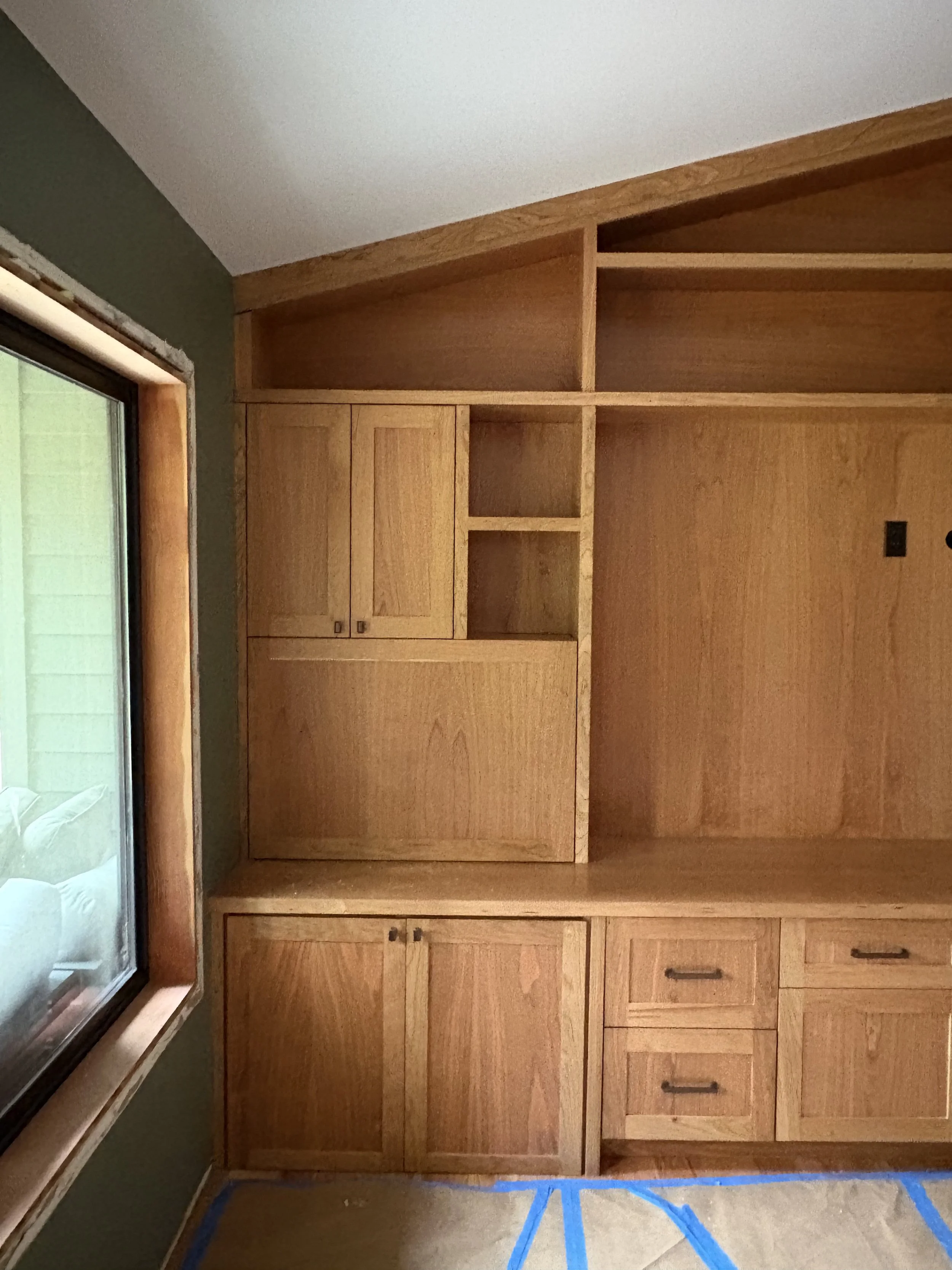 Wooden built-in cabinets and open shelving unit near a window in a room with half green wall and beige carpet.