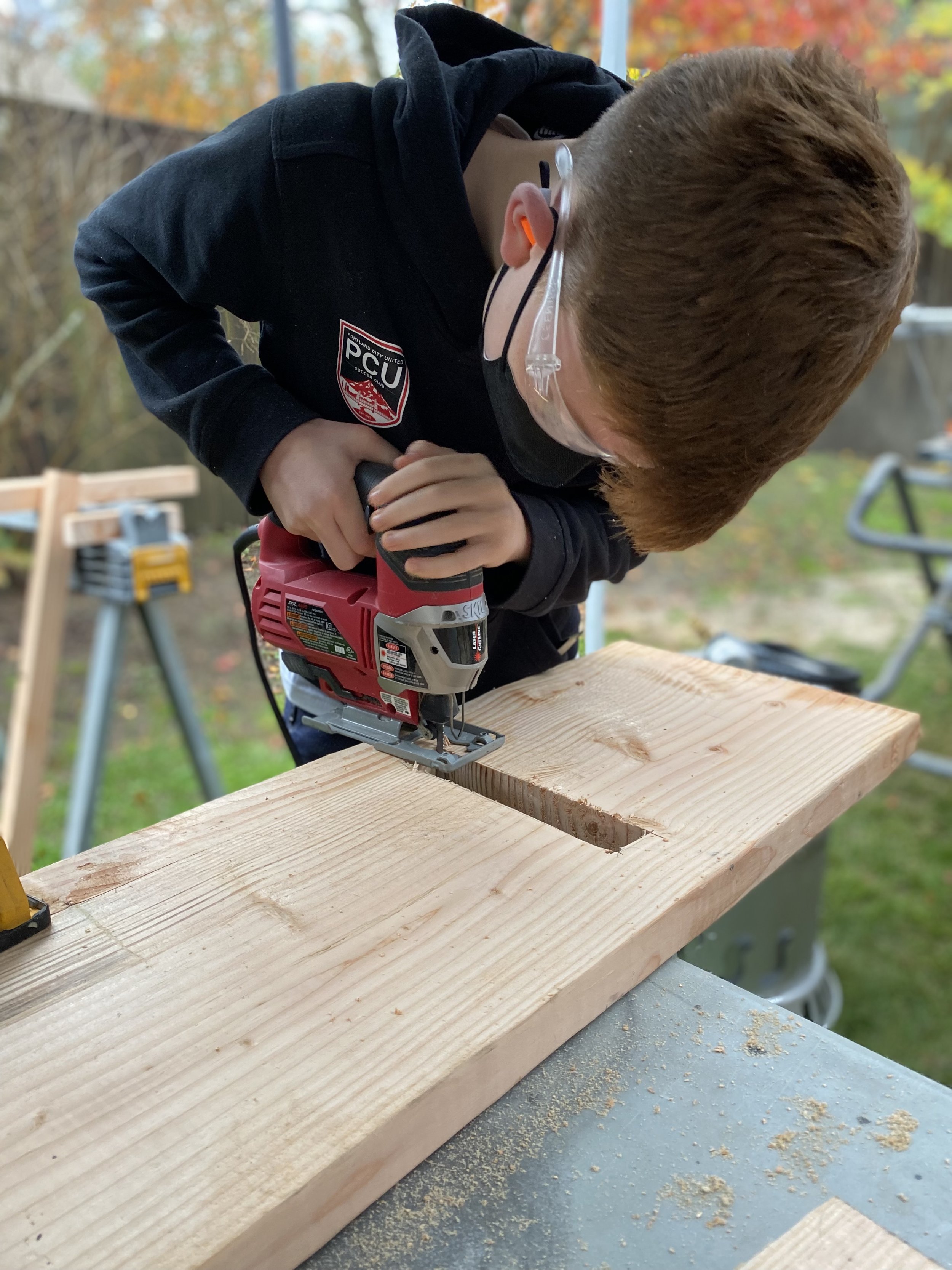 A boy wearing safety glasses and a mask uses a jigsaw to make a cut in a piece of wood outdoors.