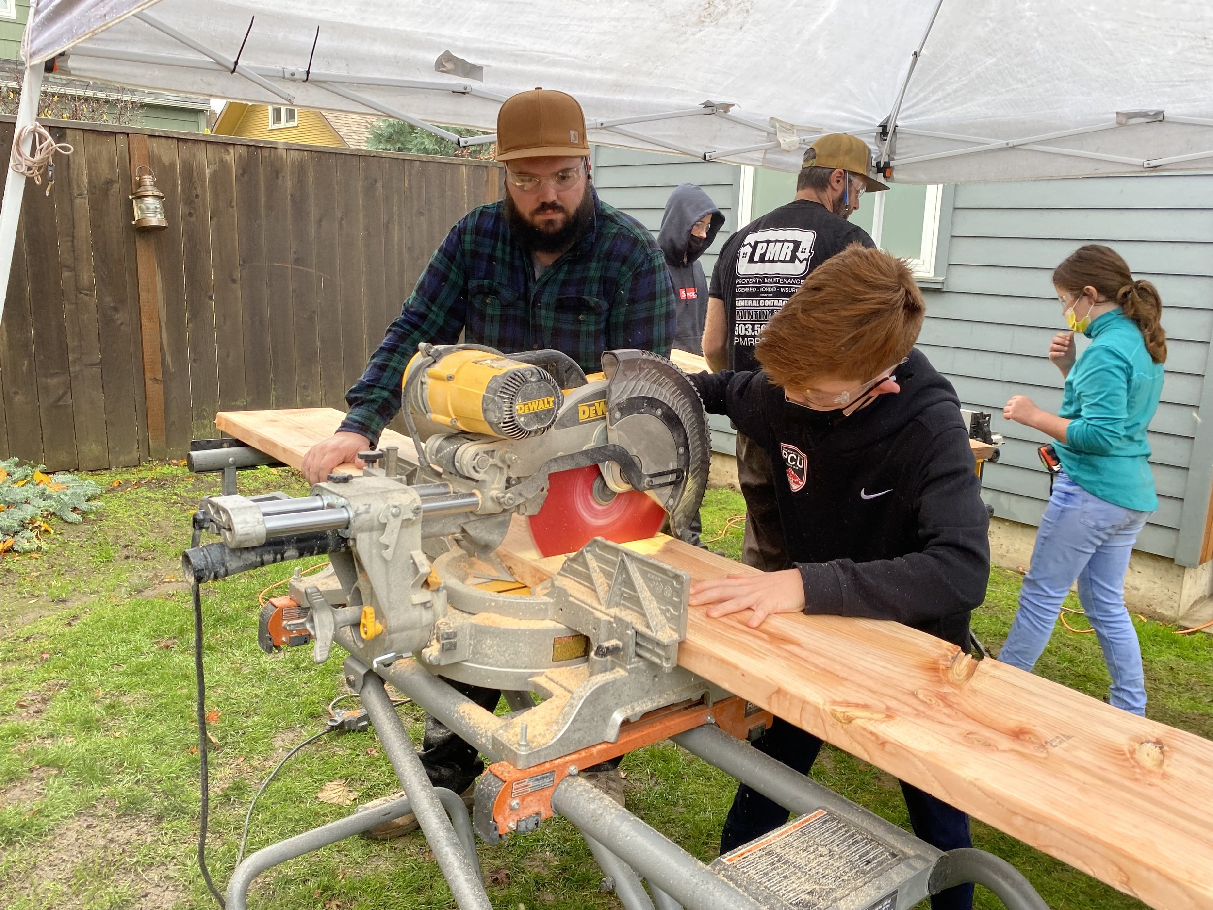 A group of people working outdoors under a canopy, with one person operating a miter saw cutting a wooden plank, while others observe or work nearby.
