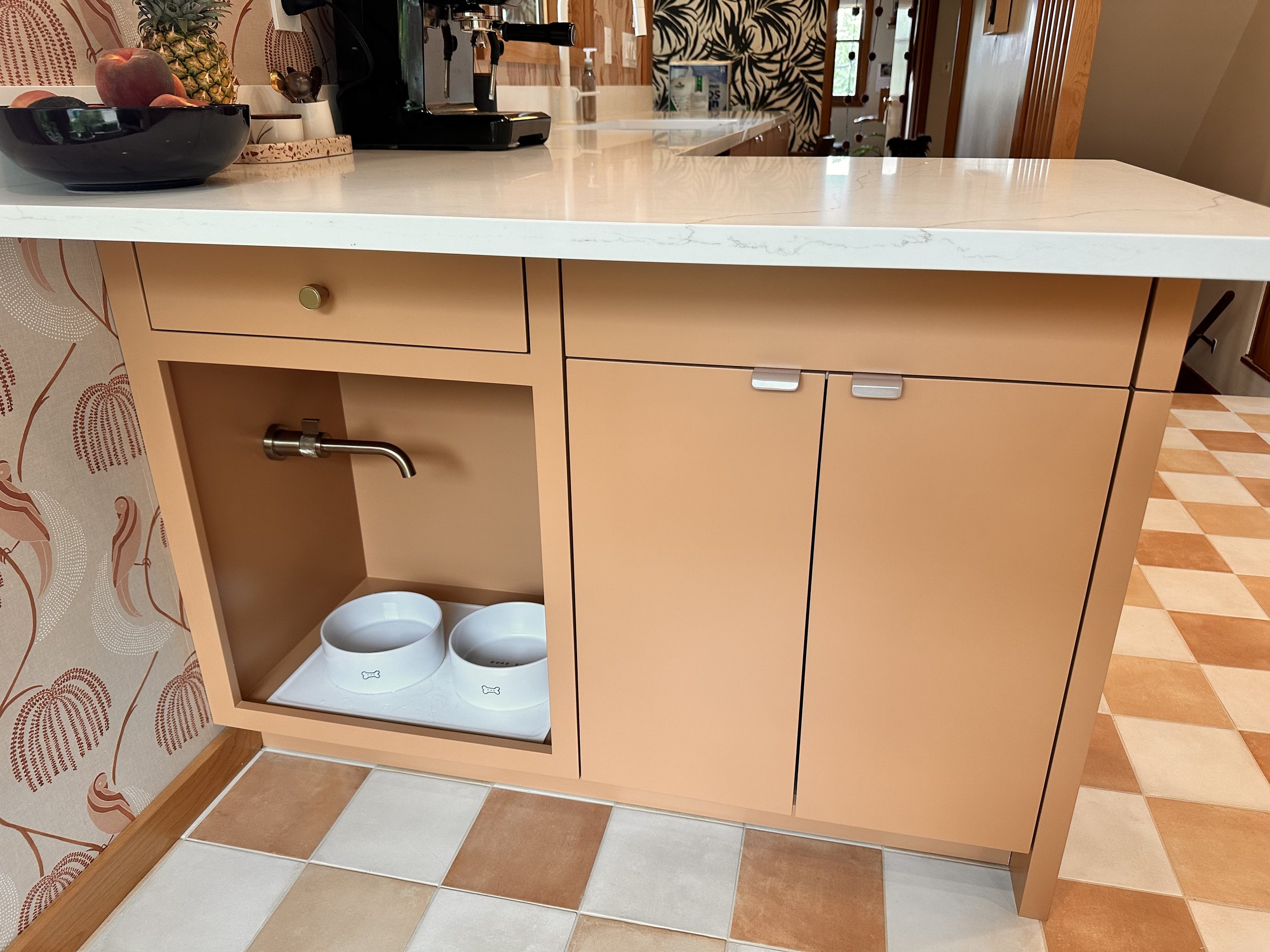 A peach-colored kitchen cabinet with a built-in pet water fountain and bowls, a white marble countertop, and various kitchen items in the background.