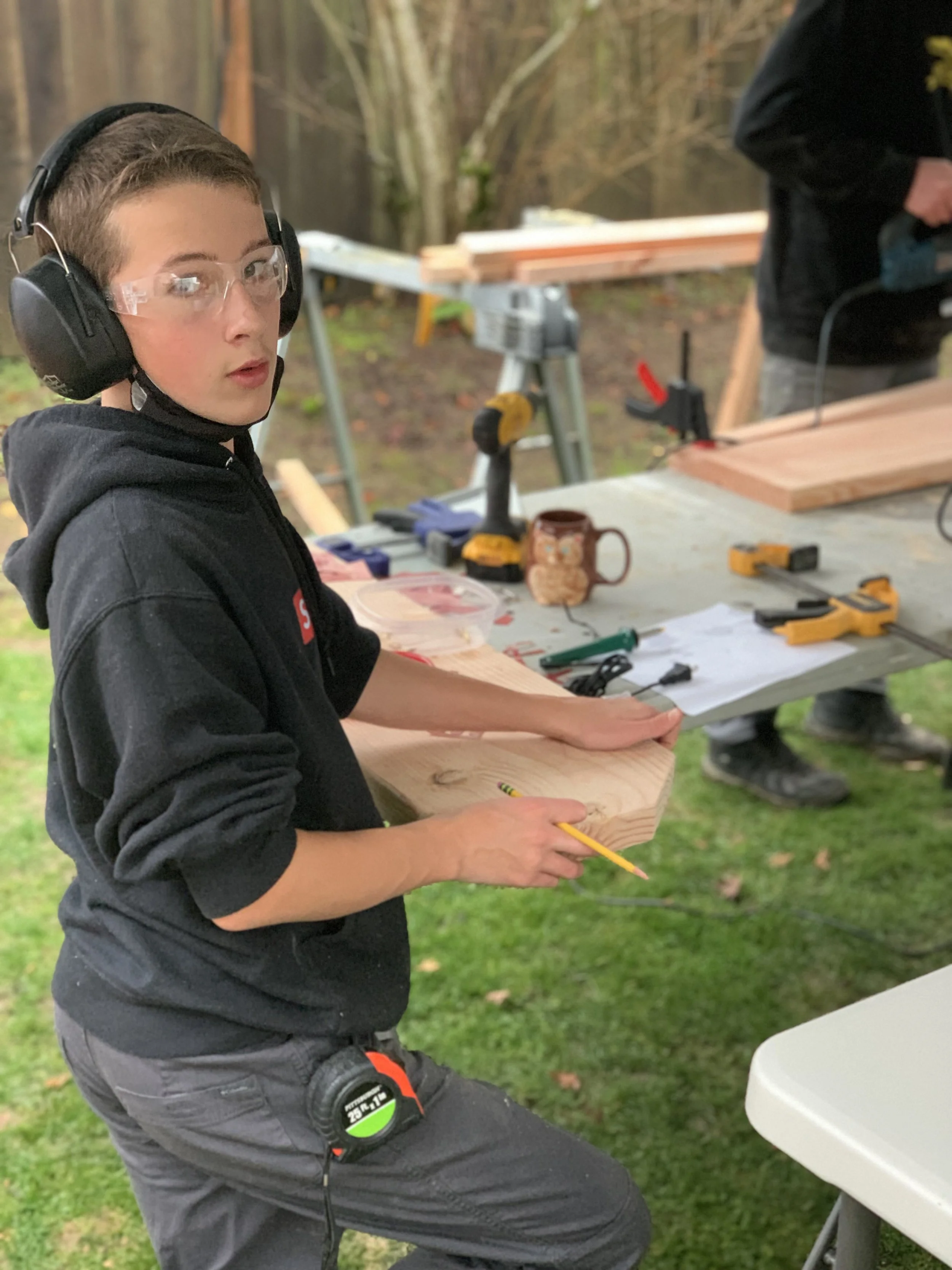 A young boy wearing safety glasses, ear protection, and a black hoodie, holding a yellow pencil while working on a wooden project at a workbench outdoors. The workspace has various tools, including a cordless drill, clamps, and a measuring tape, with