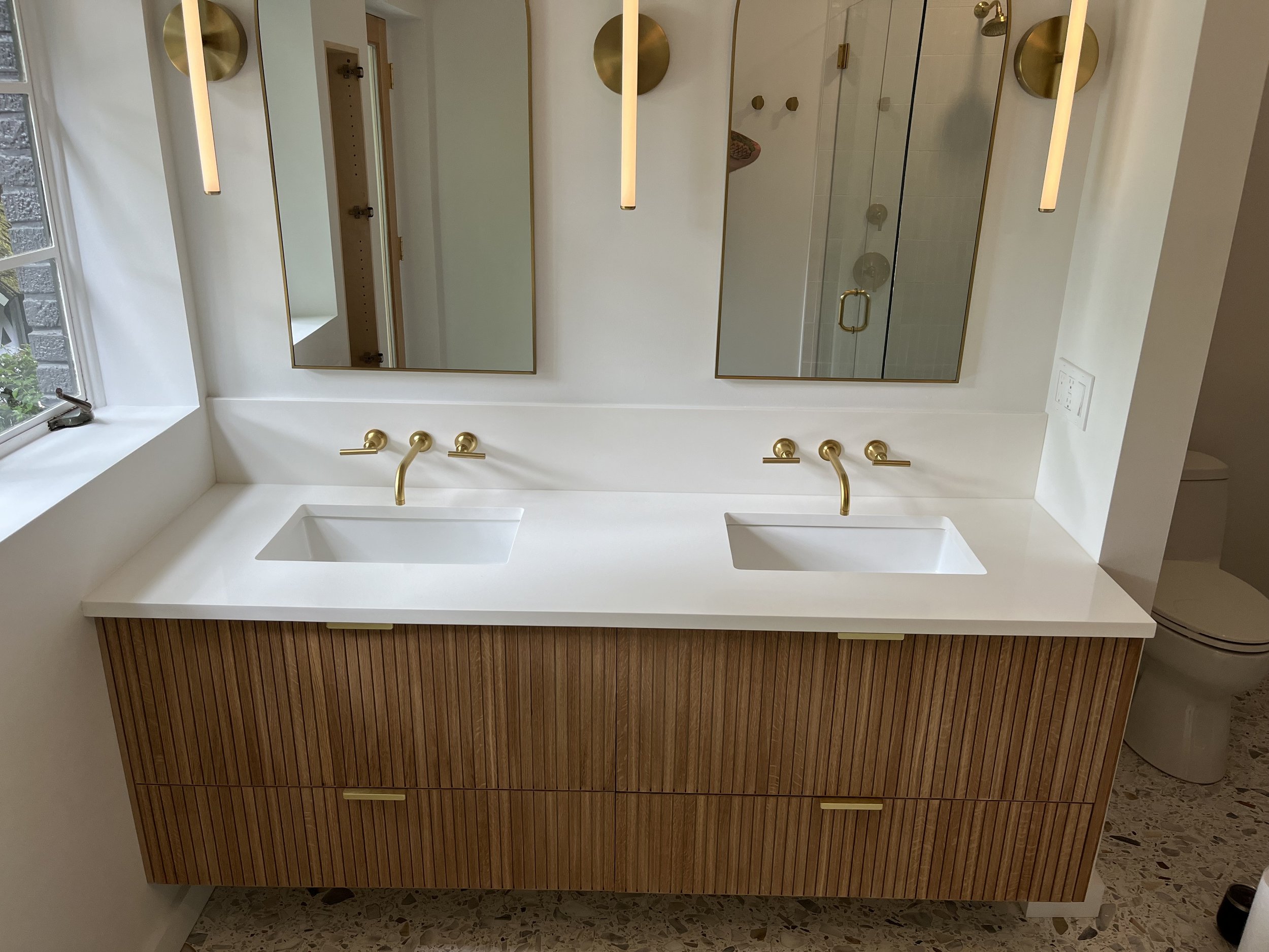 Modern dual bathroom vanity with white countertop, two rectangular sinks, gold faucets, and a large mirror. Wooden cabinet below with vertical slats and gold handles. Gold lighting fixtures and a walk-in shower with a glass door visible in the backgr