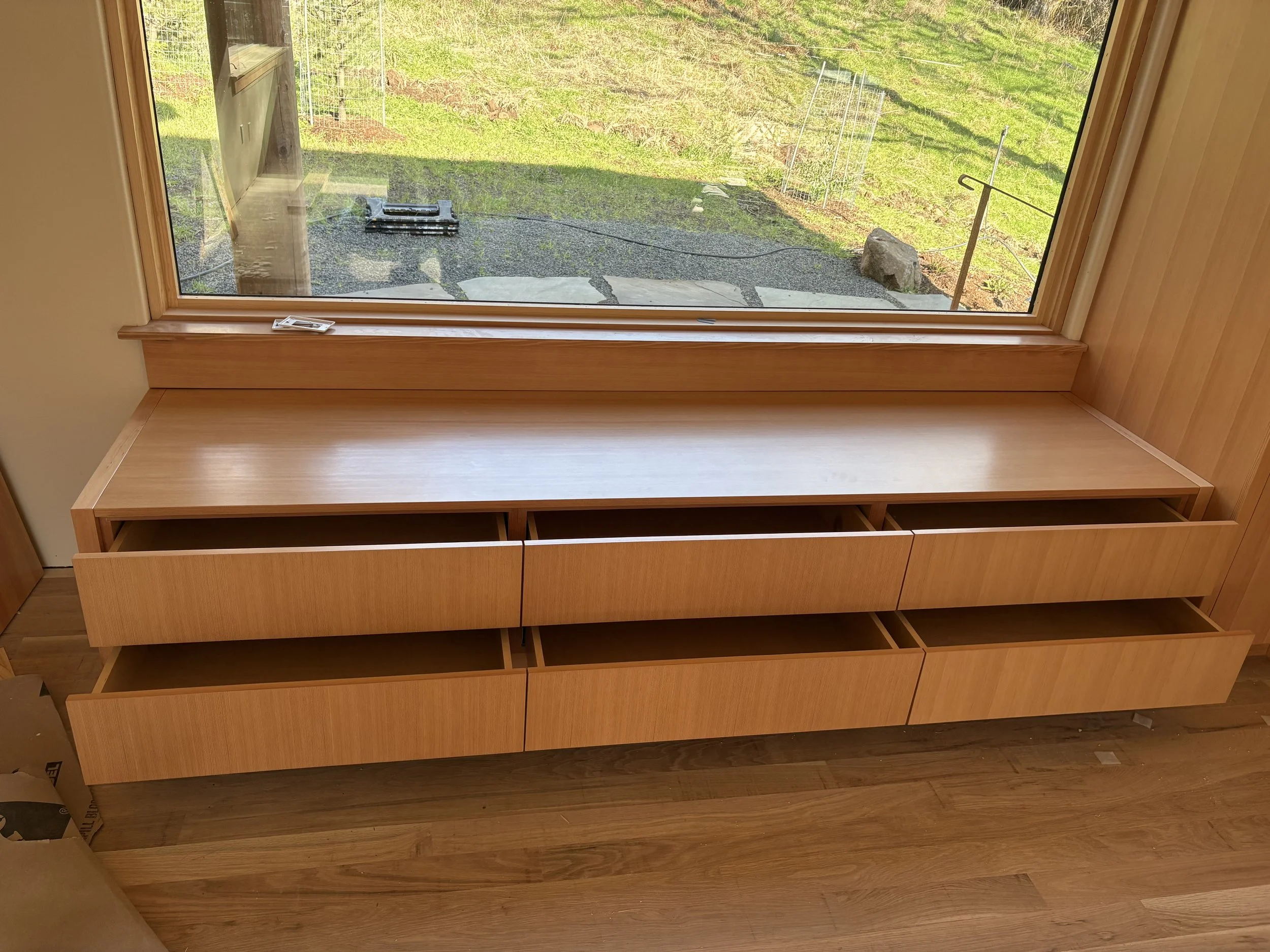 Unfinished wooden window seat with drawers underneath, positioned in front of a large window with a view of a grassy yard.