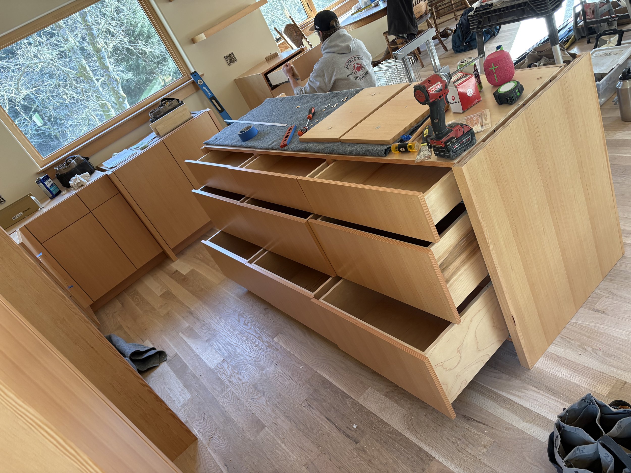 A woodworking workshop with an unfinished wooden cabinet in progress. Tools and materials such as a drill, screwdriver, tape measure, and wooden panels are on the work surface. There are open drawers in the cabinet, and a person wearing a hoodie and 