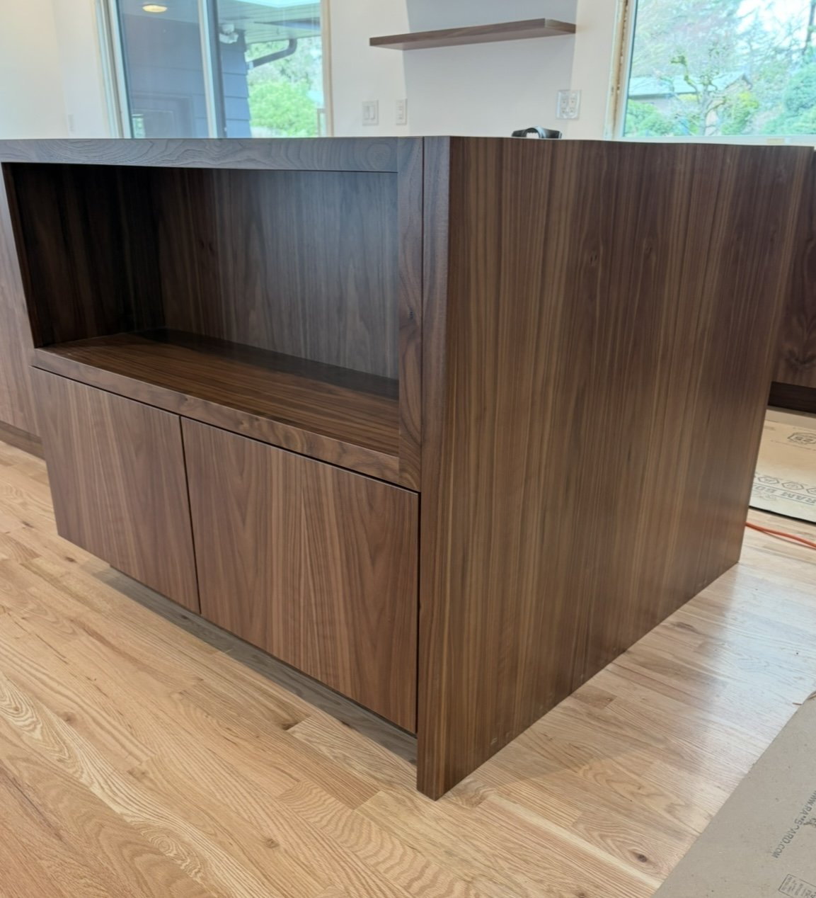A wooden cabinet with open shelving on top and closed doors below, placed on a hardwood floor inside a room with large windows and natural light.