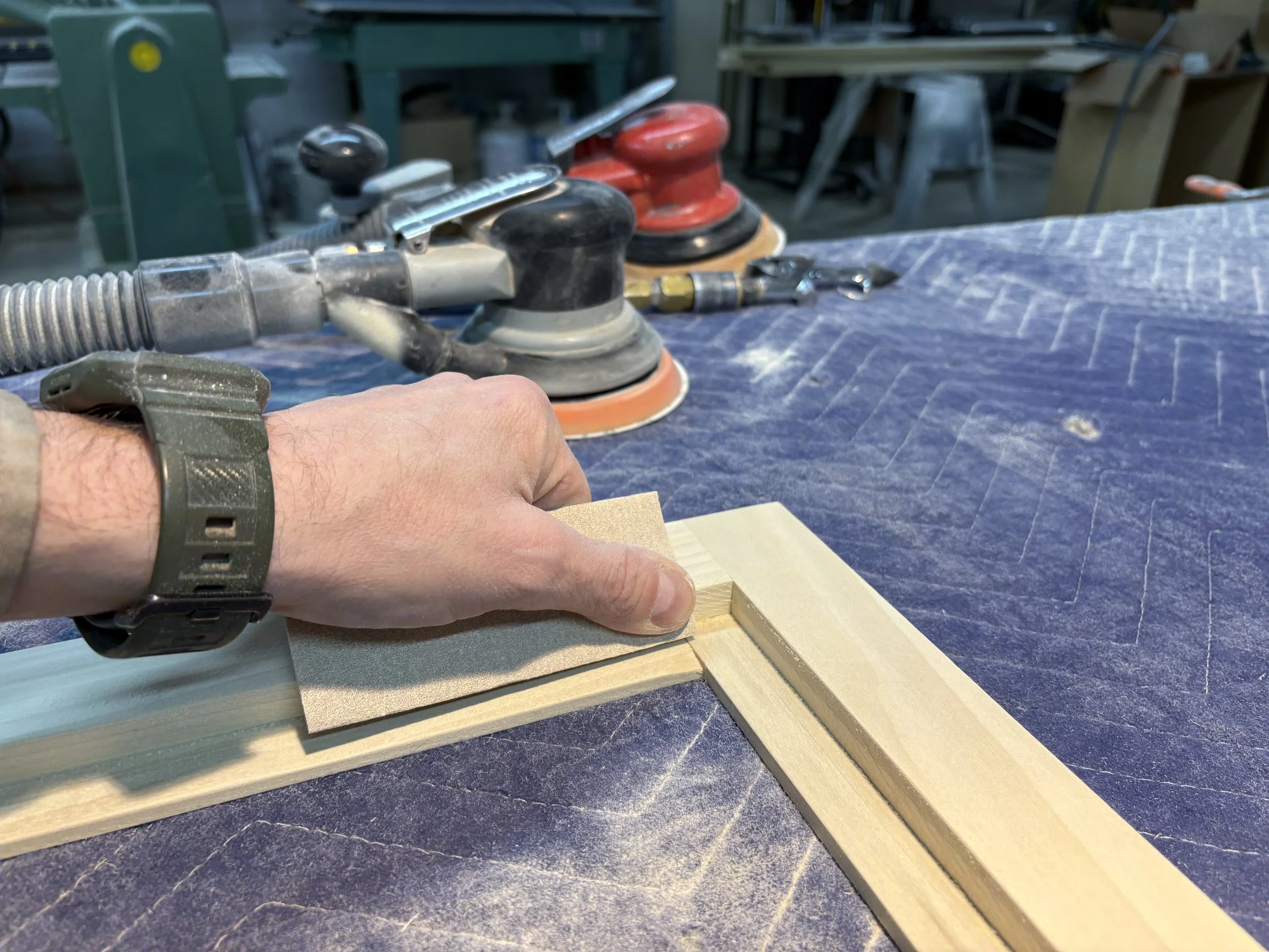 A person using a sanding tool on a piece of wood in a woodworking workshop, with various sanding and polishing machines in the background.