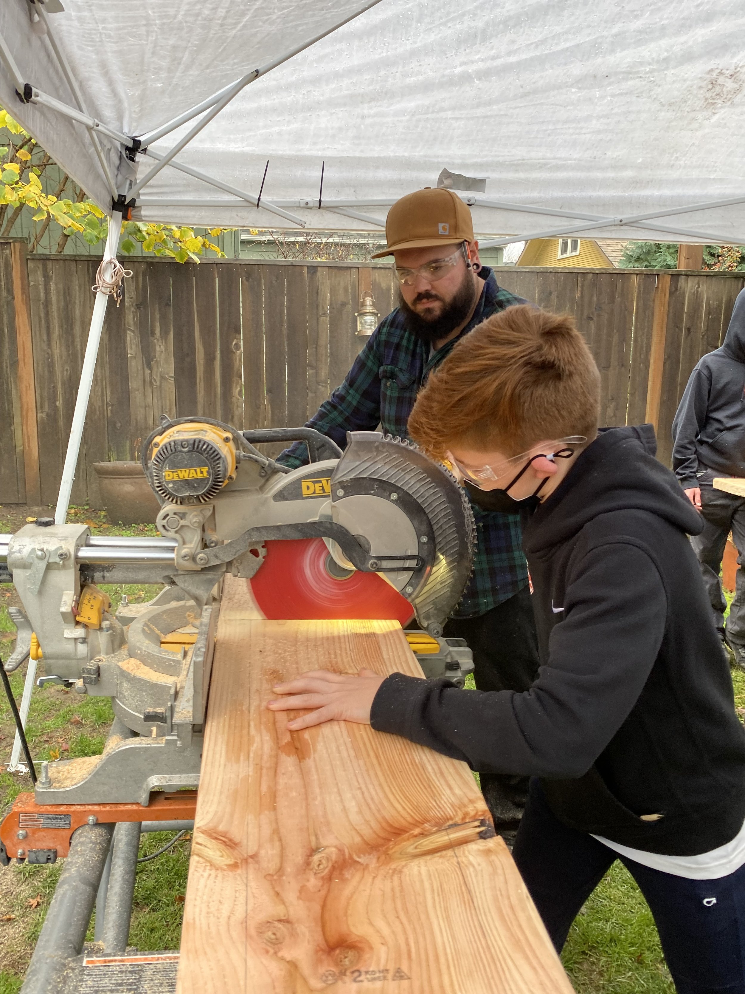 A man and a boy are cutting a wooden board with a large circular saw under a white canopy. The man is wearing glasses and a brown baseball cap, while the boy has red hair, glasses, and is wearing a black hoodie.