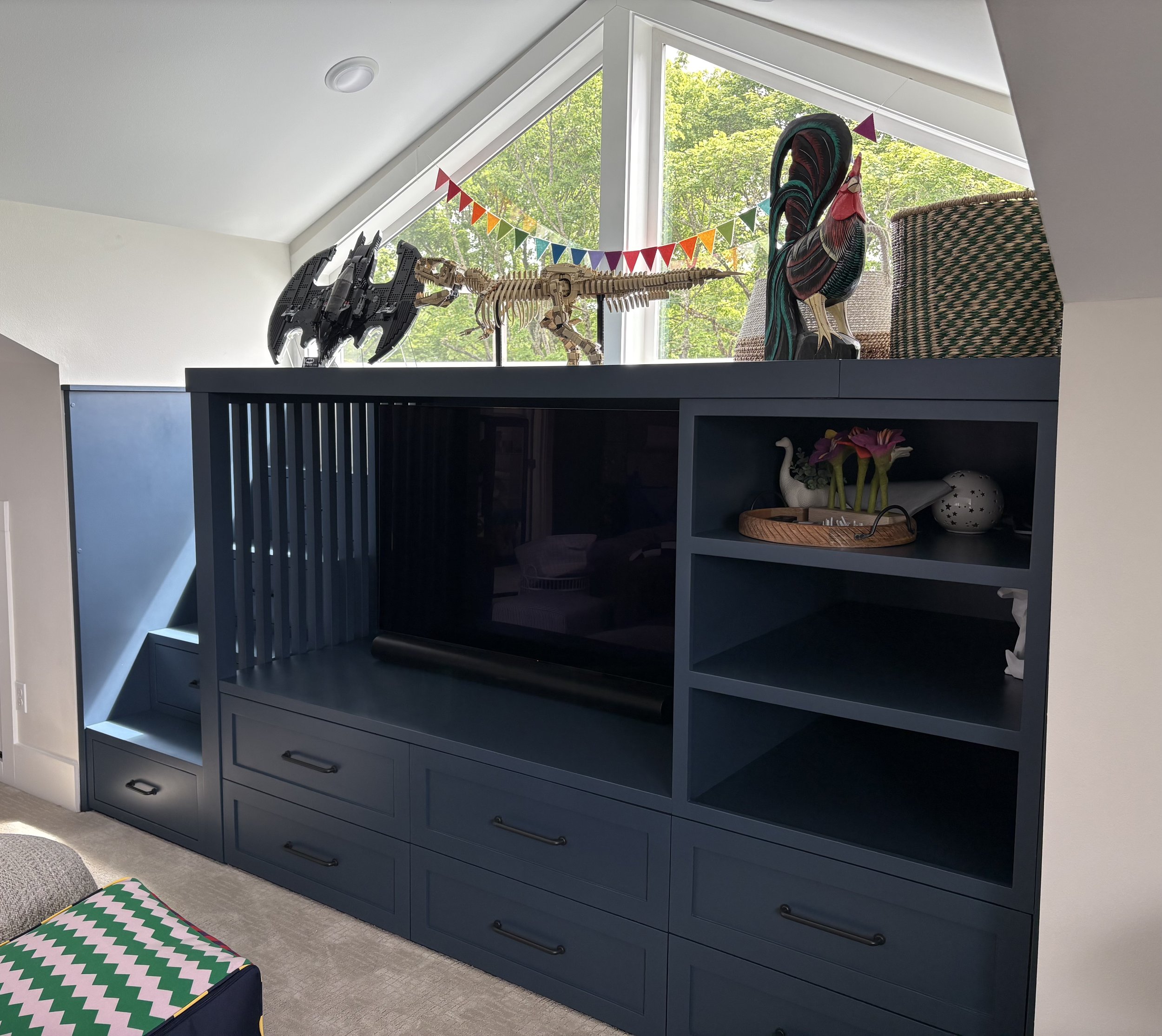 A dark blue piece of furniture with drawers and open shelves, displaying toys and decorative items, placed beneath a large window with a triangular shape. On top, there is a dinosaur model, a dragon model, and a rooster model. The window shows green 