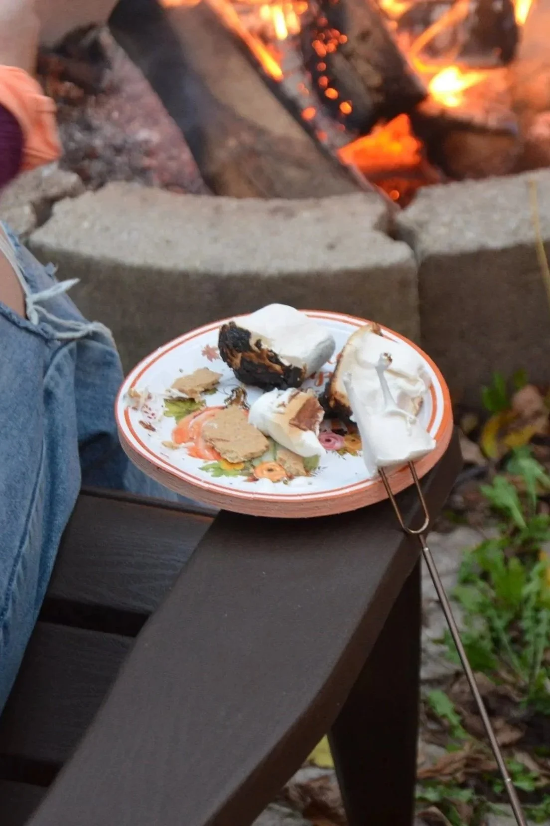 A paper plate with partially burned marshmallows and graham crackers, placed on a dark wooden table next to a person wearing ripped jeans. In the background, there is a campfire with glowing logs.