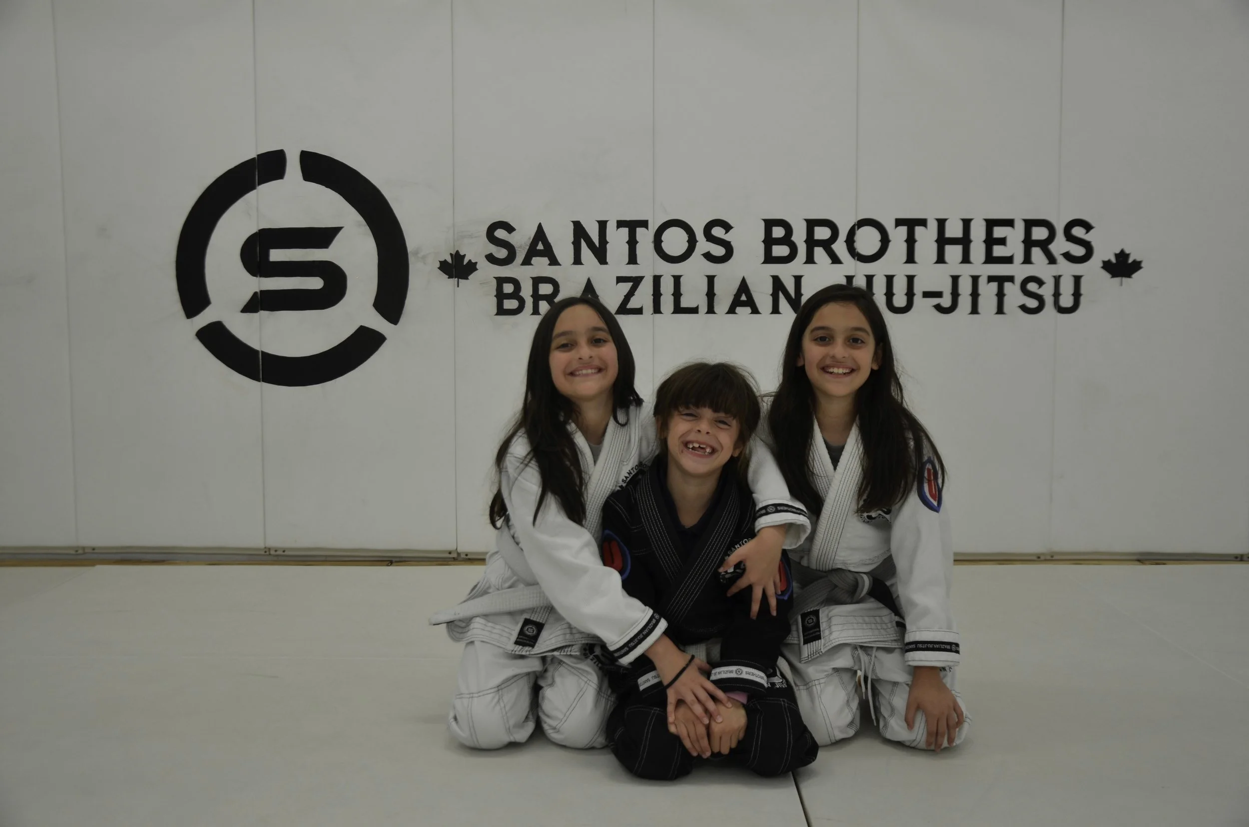 Three young girls in Brazilian Jiu-Jitsu uniforms smiling on a mat, with a white wall behind them displaying the logo and name 'Santos Brothers Brazilian Jiu-Jitsu'.