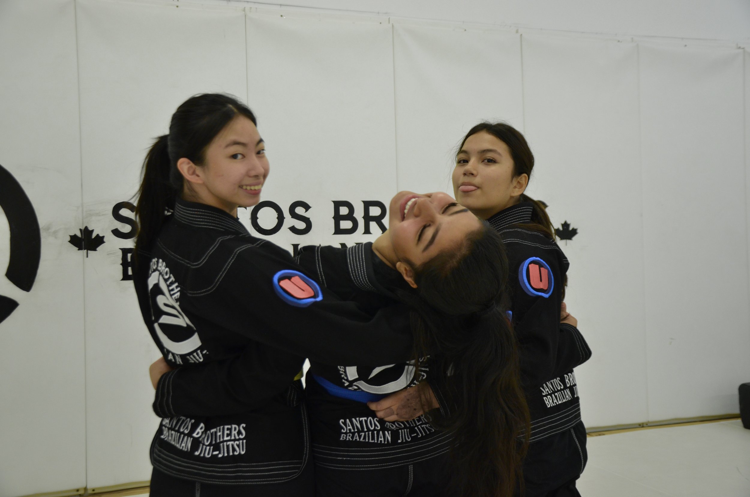 Three women in Brazilian Jiu-Jitsu uniforms holding and doing a playful pose together in a martial arts gym.