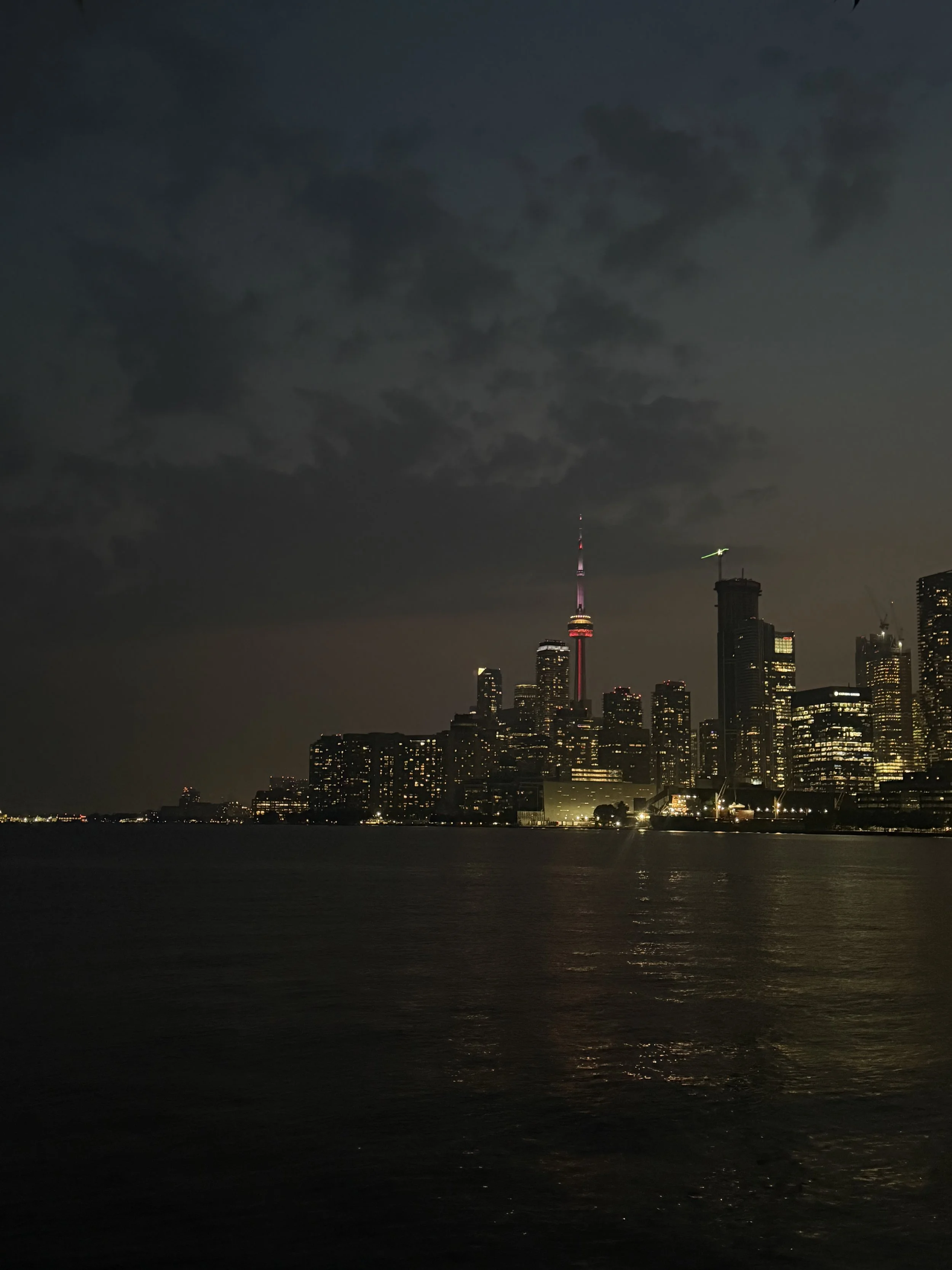 Night view of Toronto skyline with illuminated CN Tower over dark water and cloudy sky.