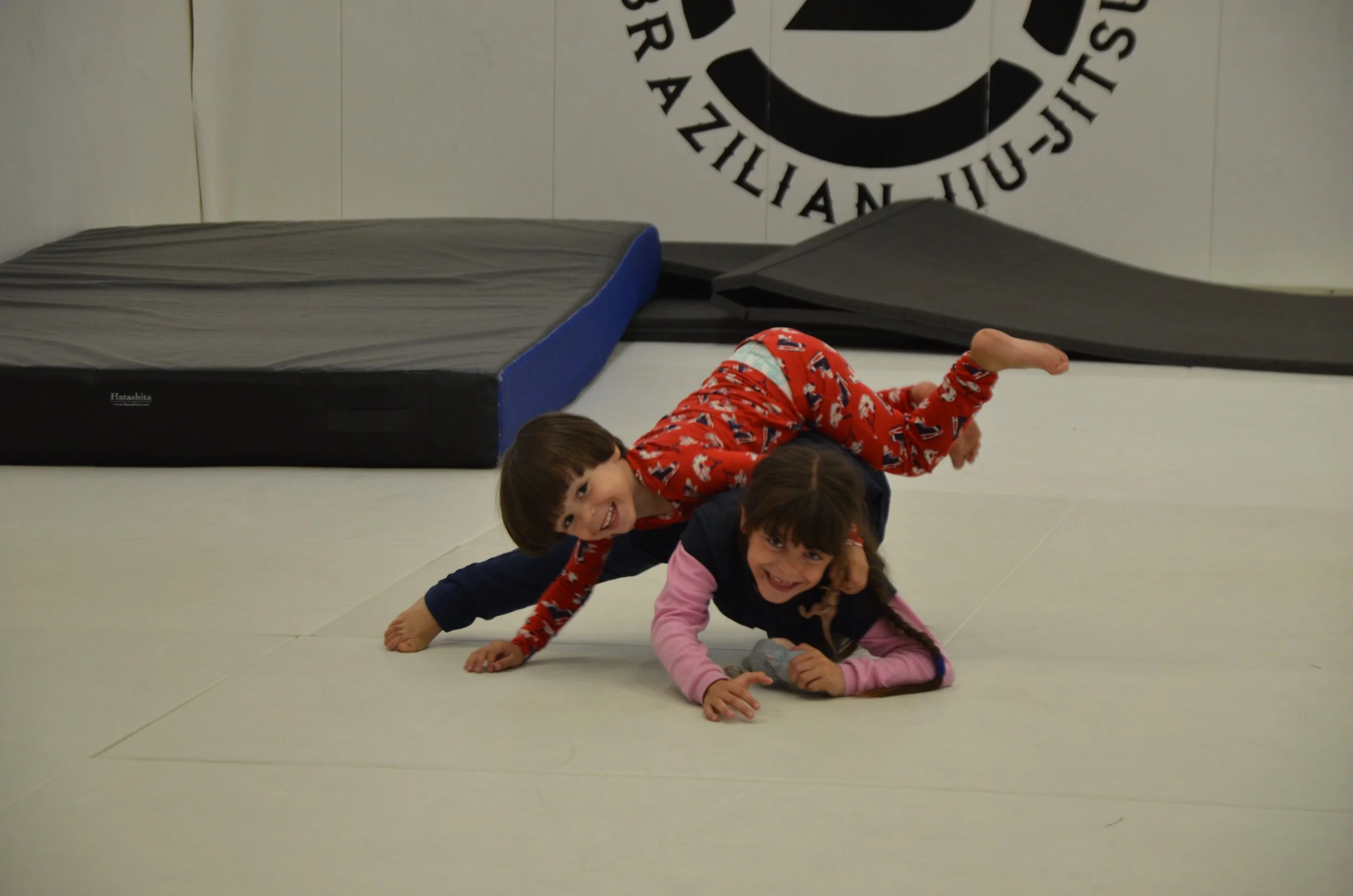 Two children playing and wrestling on a padded gym floor in a martial arts studio, with mats and a large logo on the wall behind them.
