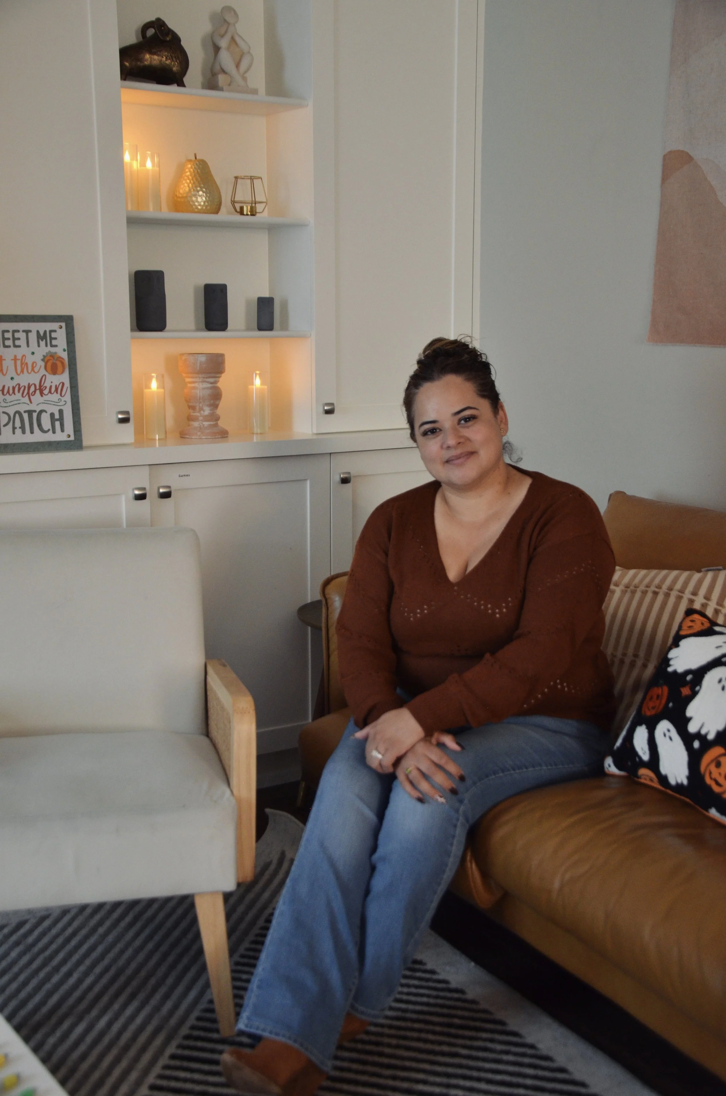 A woman sitting on a tan leather couch in a living room with decorated shelves and pillows.