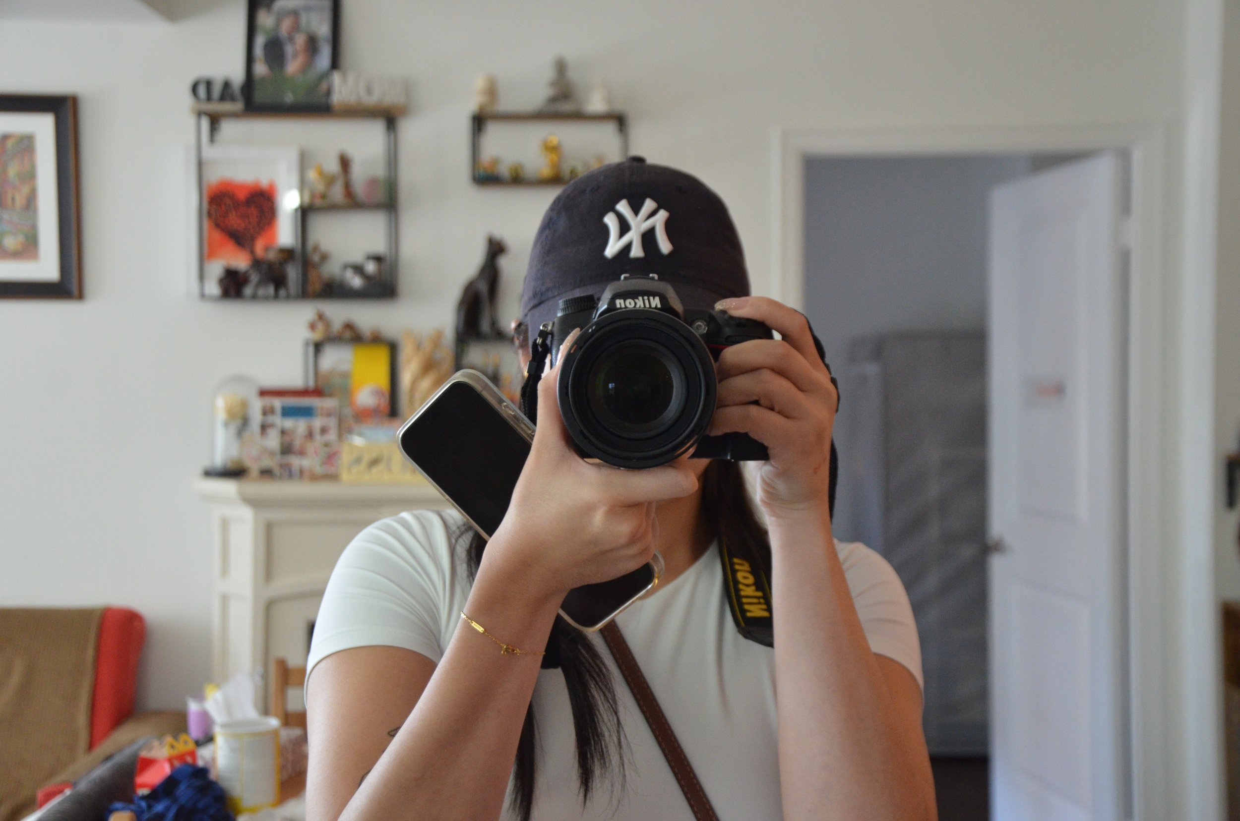 Person taking a photo in front of a mirror with a camera, wearing a baseball cap and a white t-shirt, holding a phone in their hand.