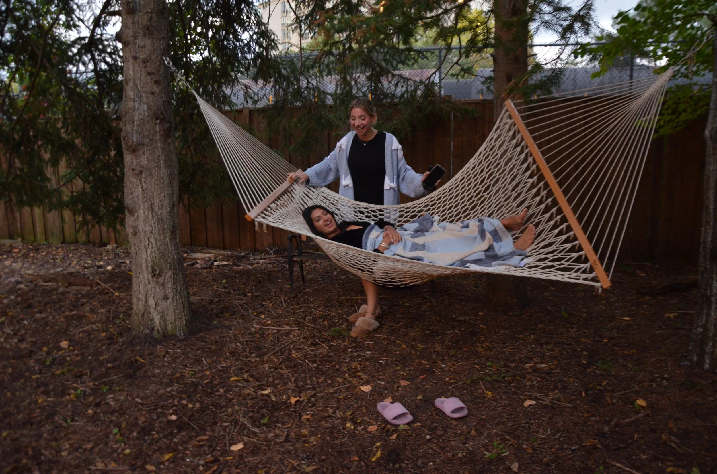 Two women relaxing and laughing in a hammock in a backyard, with one standing and the other lying down wrapped in a blanket, surrounded by trees and a wooden fence.