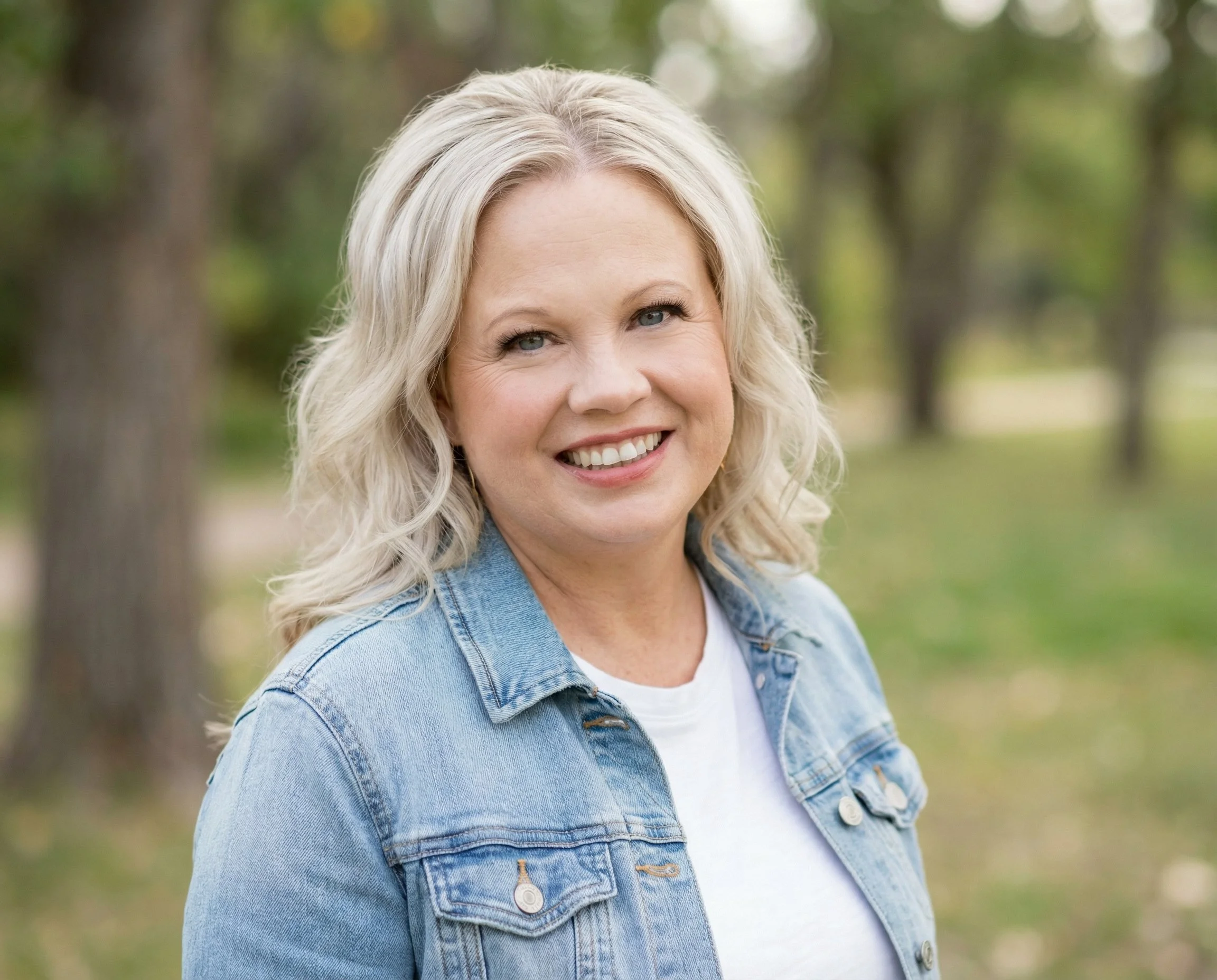 A smiling blonde woman wearing a denim jacket outdoors with trees in the background.