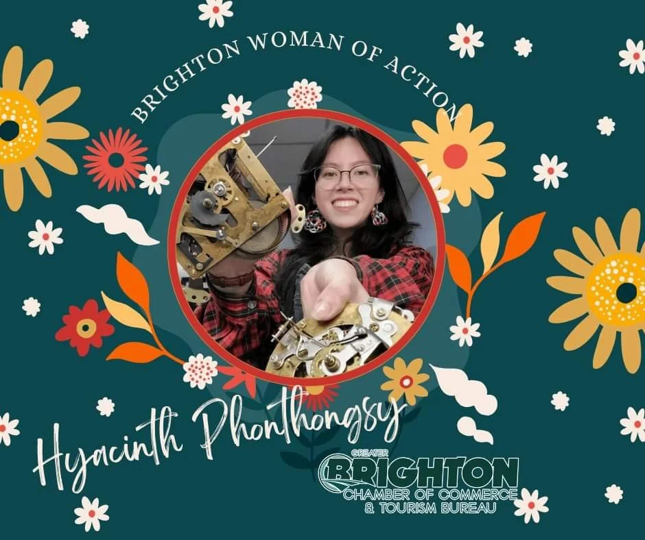 The clockmaker smiling and holding mechanical clock parts, surrounded by text naming her a Brighton Woman of Action, with the Greater Brighton Chamber of Commerce and Tourism Bureau logo.