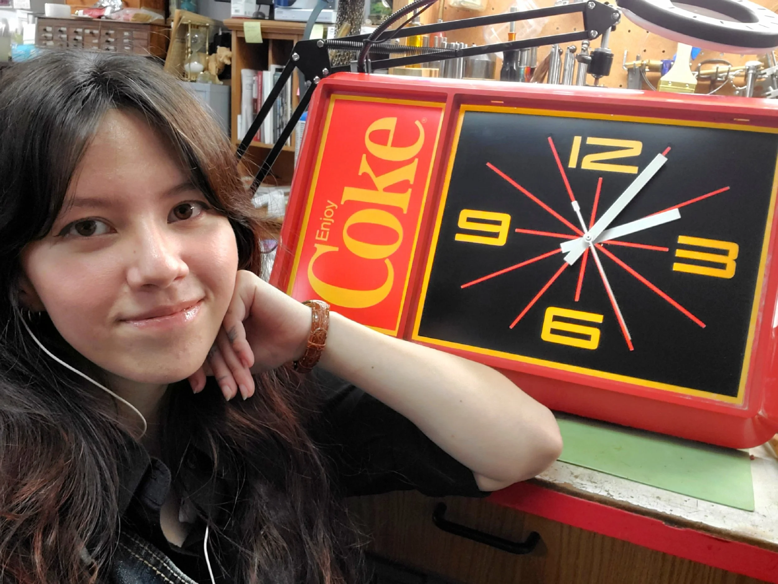 A woman with long dark hair is smiling with her head resting on her hand. She is sitting next to a vintage electric Coca-Cola clock with a black face, yellow numbers, and red details in a workshop with tools in the background.