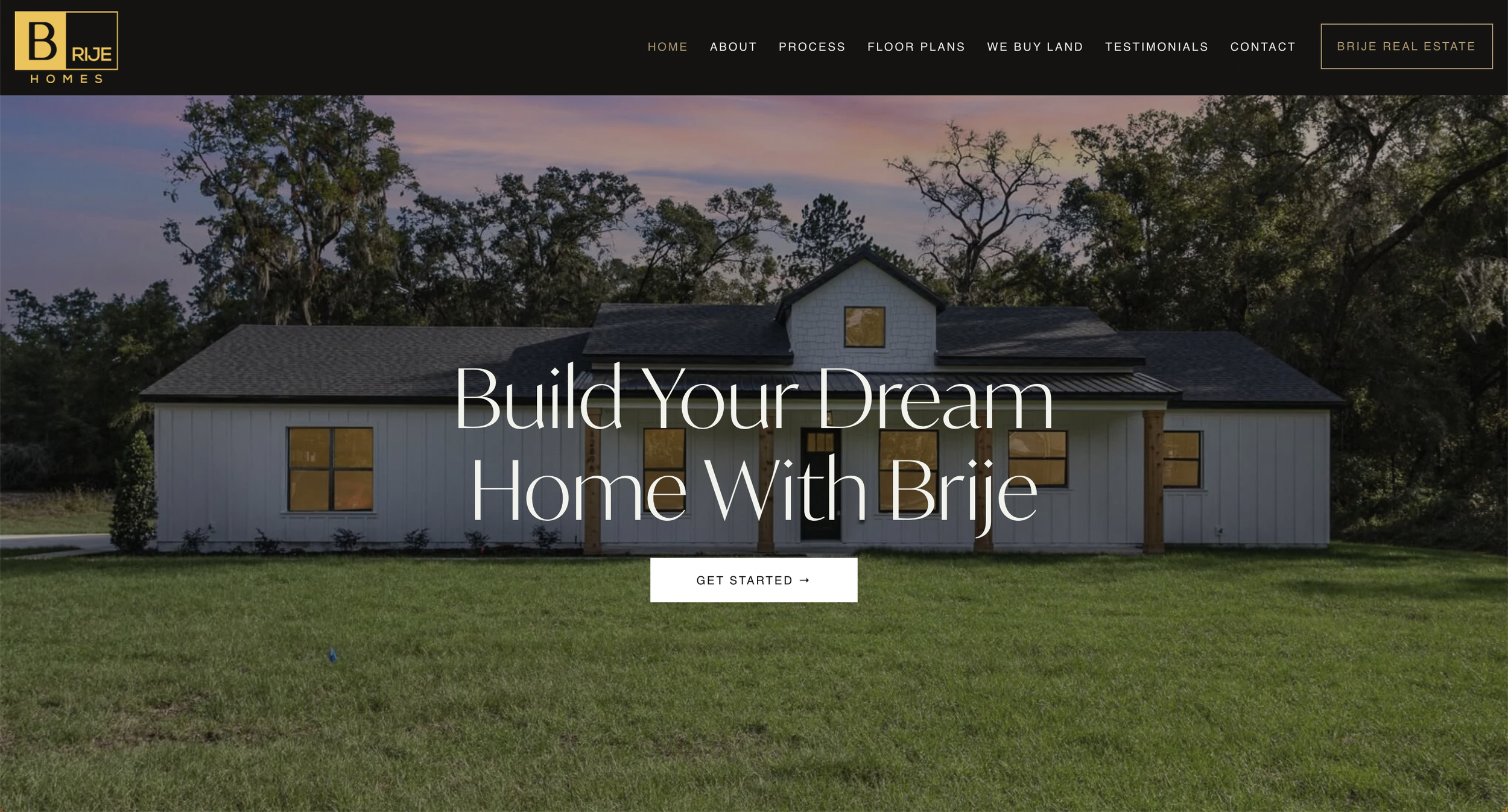 Front view of a newly built white house with a dark roof, surrounded by a grassy yard and trees in the background, at sunset.