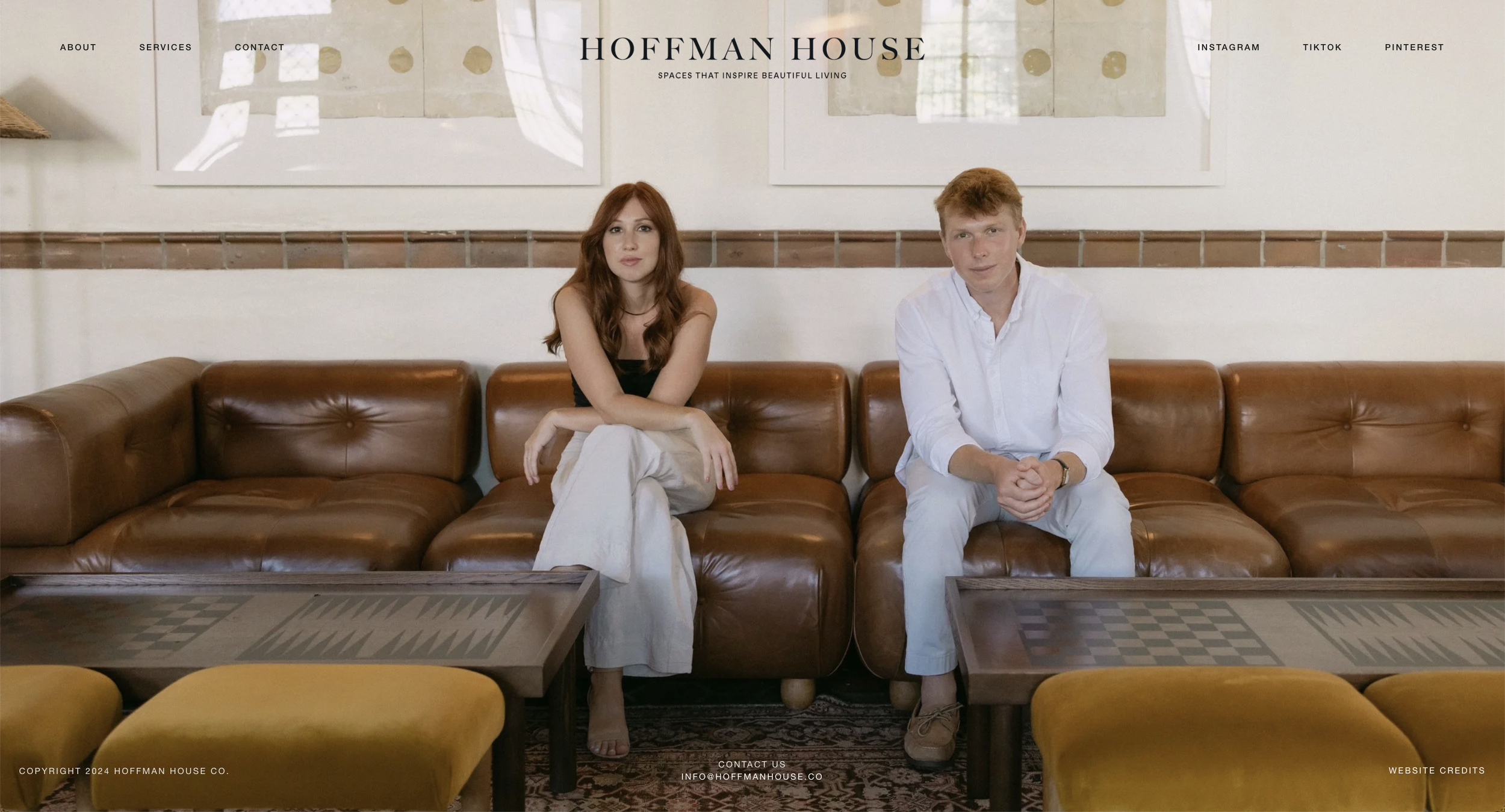 A woman and a man sit on a brown leather couch in a modern living room. The woman has long red hair and is wearing a black top and white pants. The man has short blond hair and is wearing a white shirt and light-colored pants. Behind them are two framed abstract art pieces on the wall, with a logo that reads 'Hoffman House' and a tagline 'Spaces That Inspire Beautiful Living.'