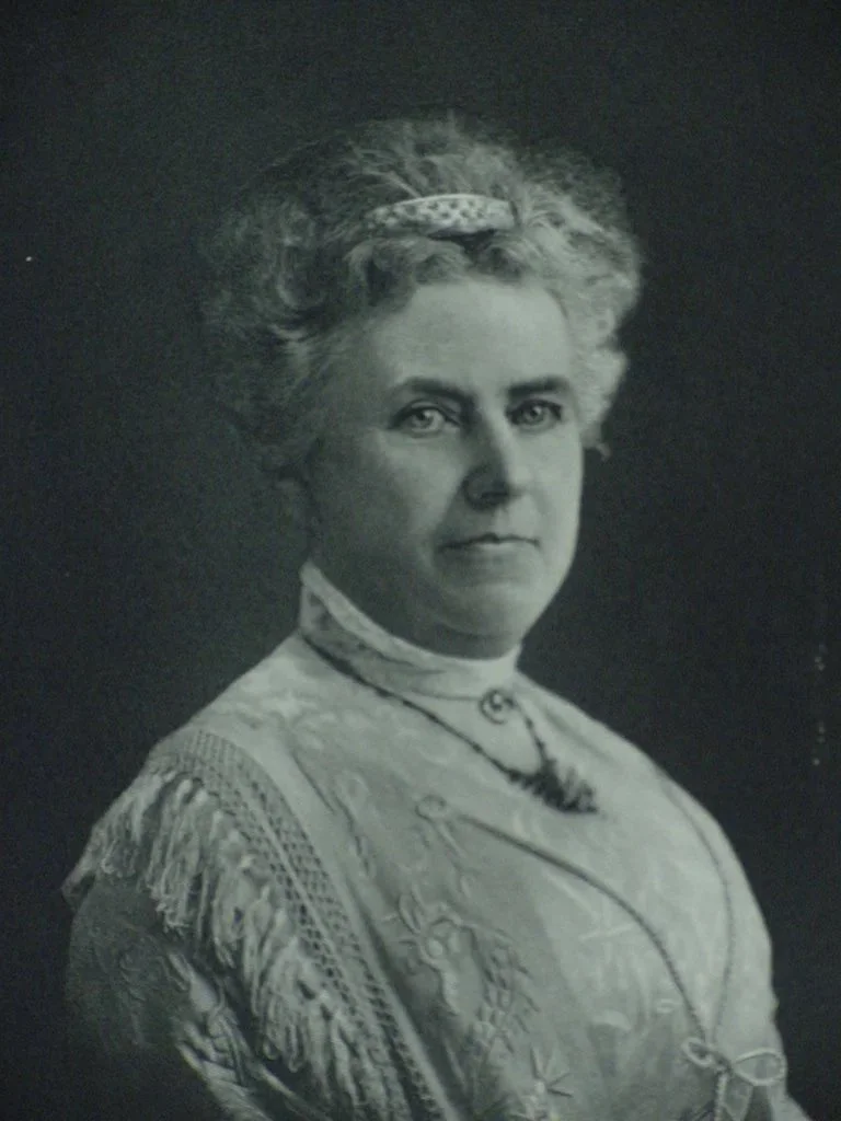 Black and white portrait of a woman with gray, curly hair wearing a patterned dress, a necklace, and a small hat.