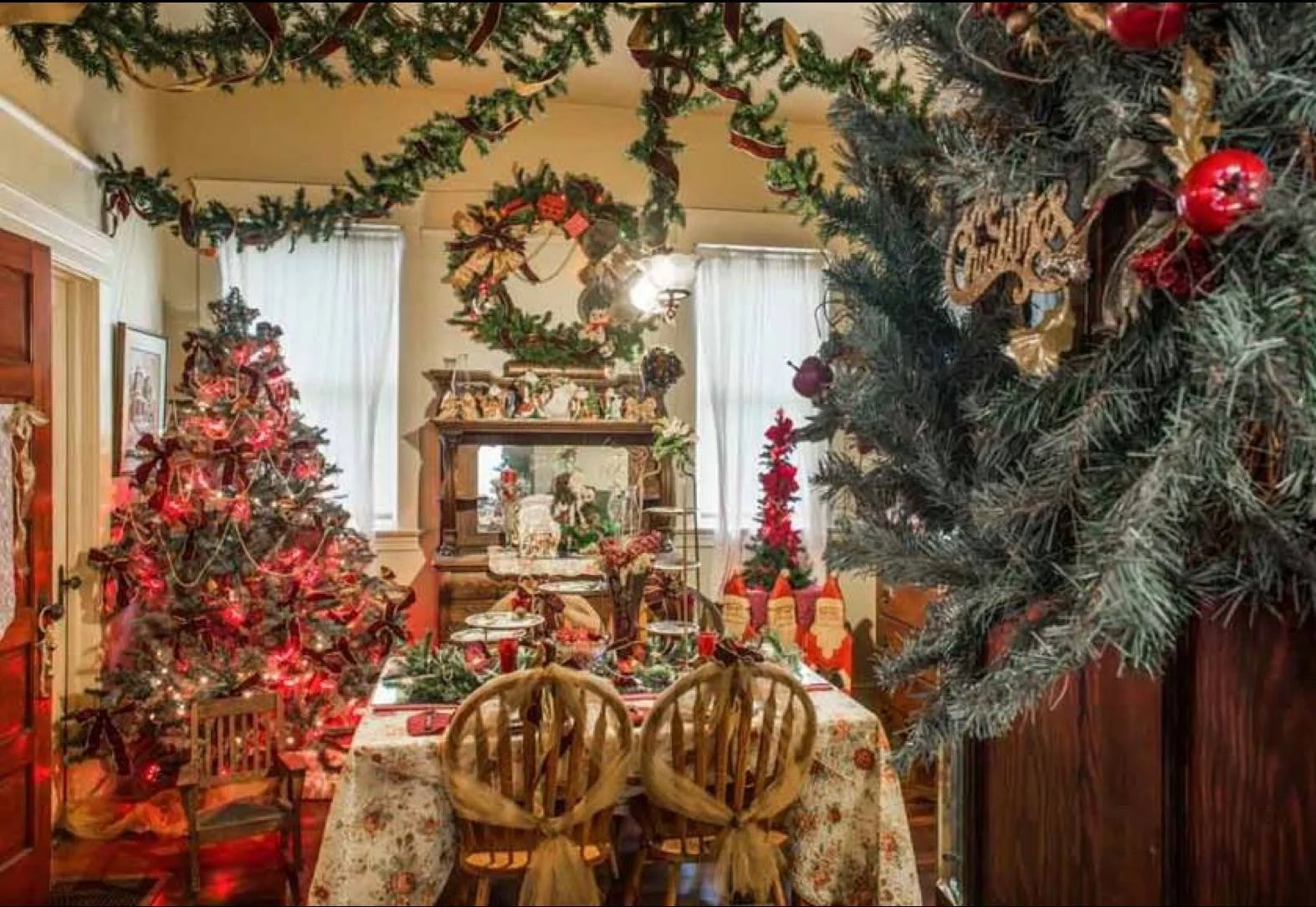 Festively decorated dining room with Christmas trees, garlands, and stockings. The table is covered with a holiday tablecloth and set for a meal, with chairs tied with ribbons. The room has large windows with white curtains and a wooden china cabinet.