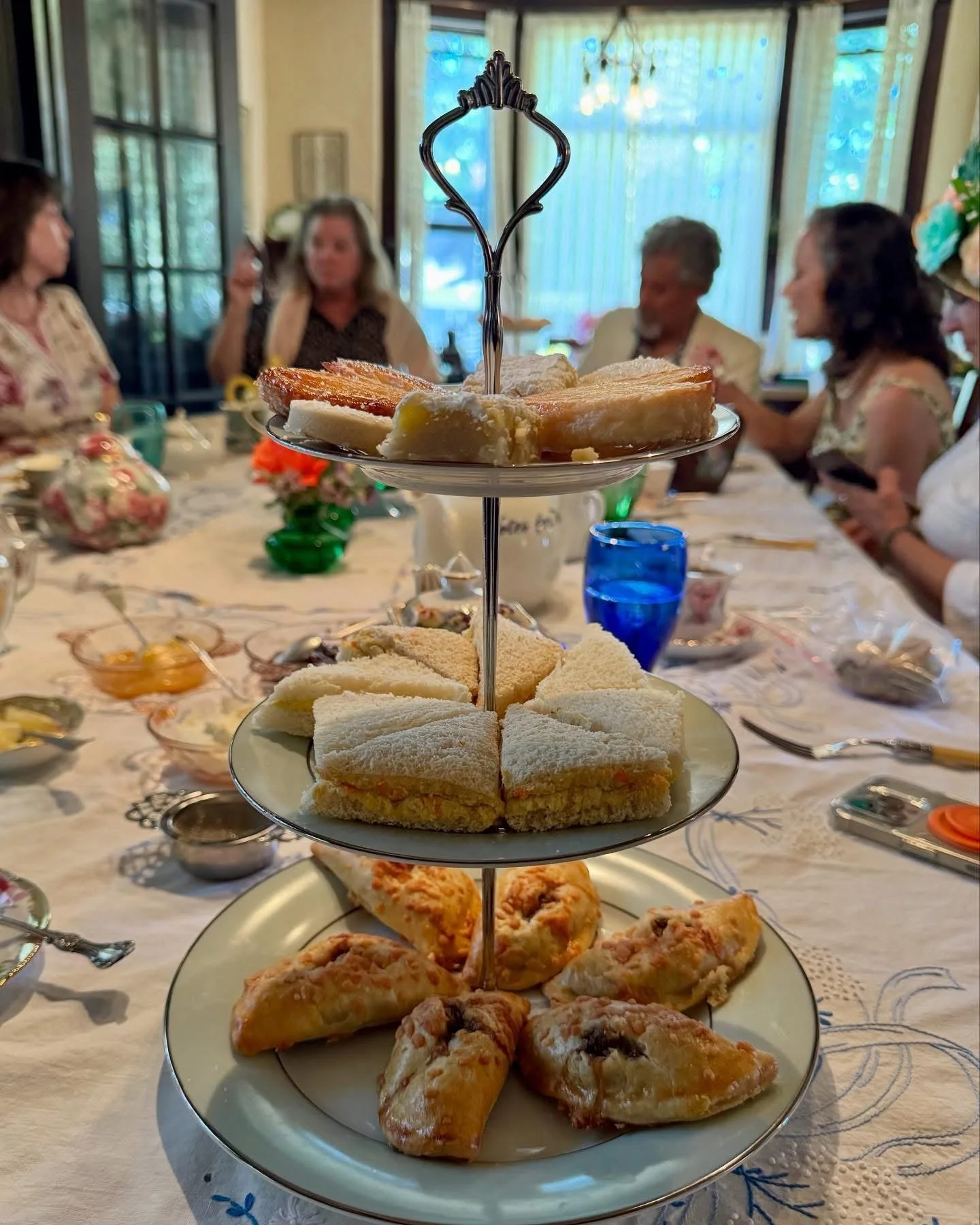 A three-tiered serving tray with assorted sandwiches, scones, and small pastries at a tea party, with women seated at a decorated table in the background.