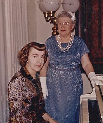 Two women, one younger, Dorothy Bembridge with curly hair sitting at a piano and an older woman, unknown, standing beside her, wearing a blue dress, glasses, and pearl jewelry, in a living room with curtains and a lamp.