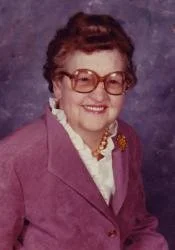 A portrait of an elderly woman with short, curly hair and glasses, wearing a purple blazer with a white blouse and a brooch, smiling against a plain background.