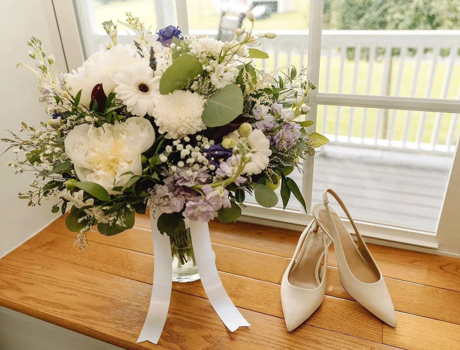 A bouquet of white and purple flowers in a glass vase, paired with a pair of white high-heeled shoes on a wooden surface near a window.