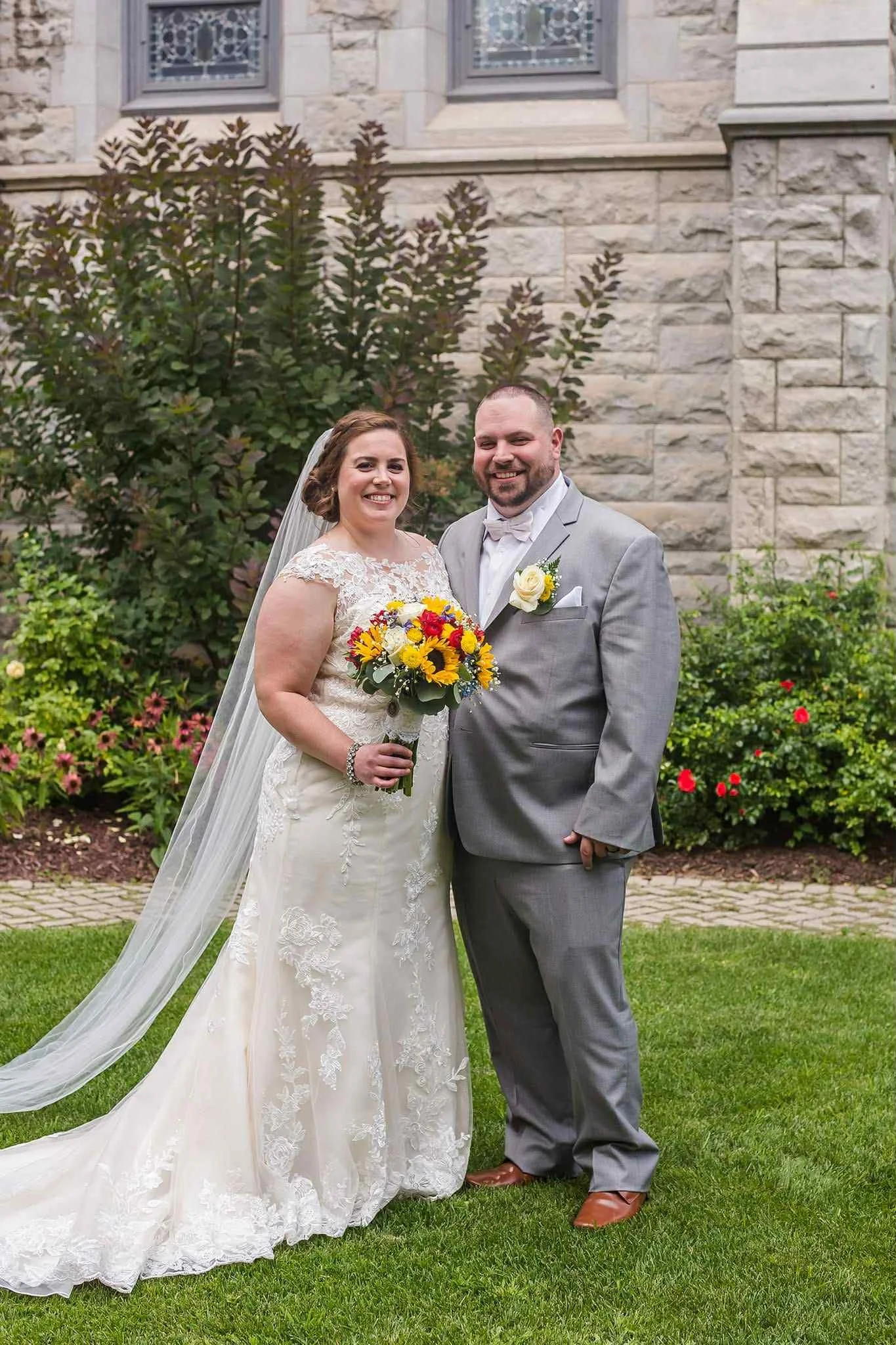 A bride and groom standing outdoors in front of a stone building and flowering bushes, smiling for a wedding photo. The bride is holding a bouquet of yellow, red, and white flowers and is wearing a lace wedding gown with a veil. The groom is dressed 