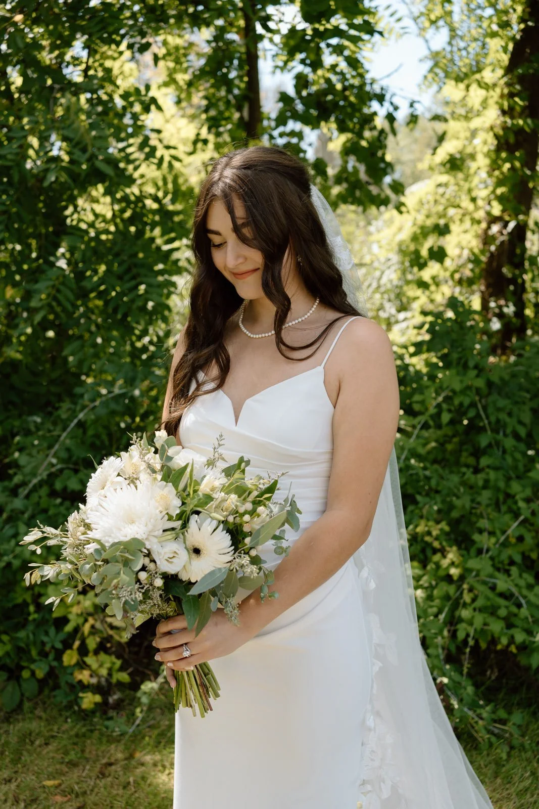 A bride in a white wedding dress holding a bouquet of white flowers in an outdoor setting with green foliage.