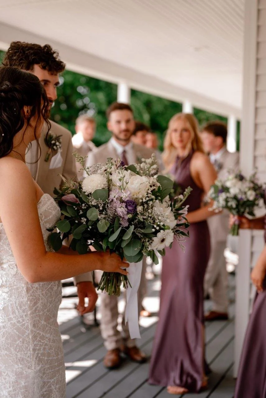 A bride holding a bouquet of white and purple flowers at her wedding, with several guests and bridesmaids in formal attire in the background.