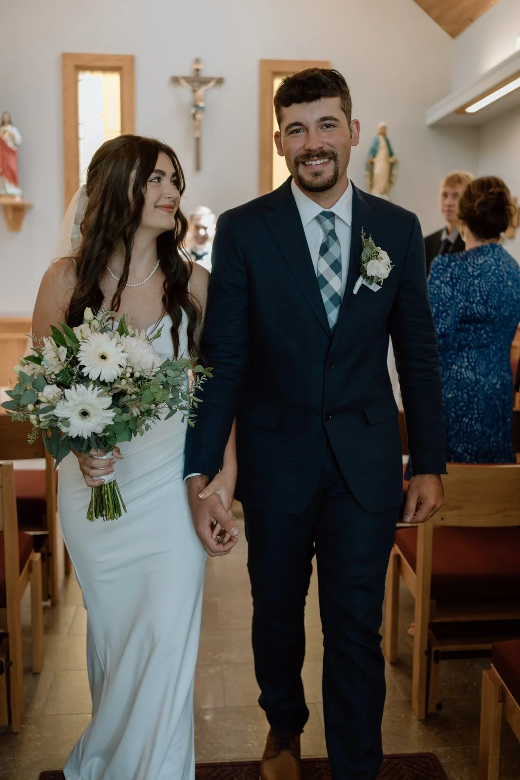 A bride and groom walking down the aisle in a church, holding hands. The bride has long dark hair and wears a white wedding dress with a bouquet of white flowers. The groom has short dark hair and a beard, wearing a dark suit with a tie, and a white 