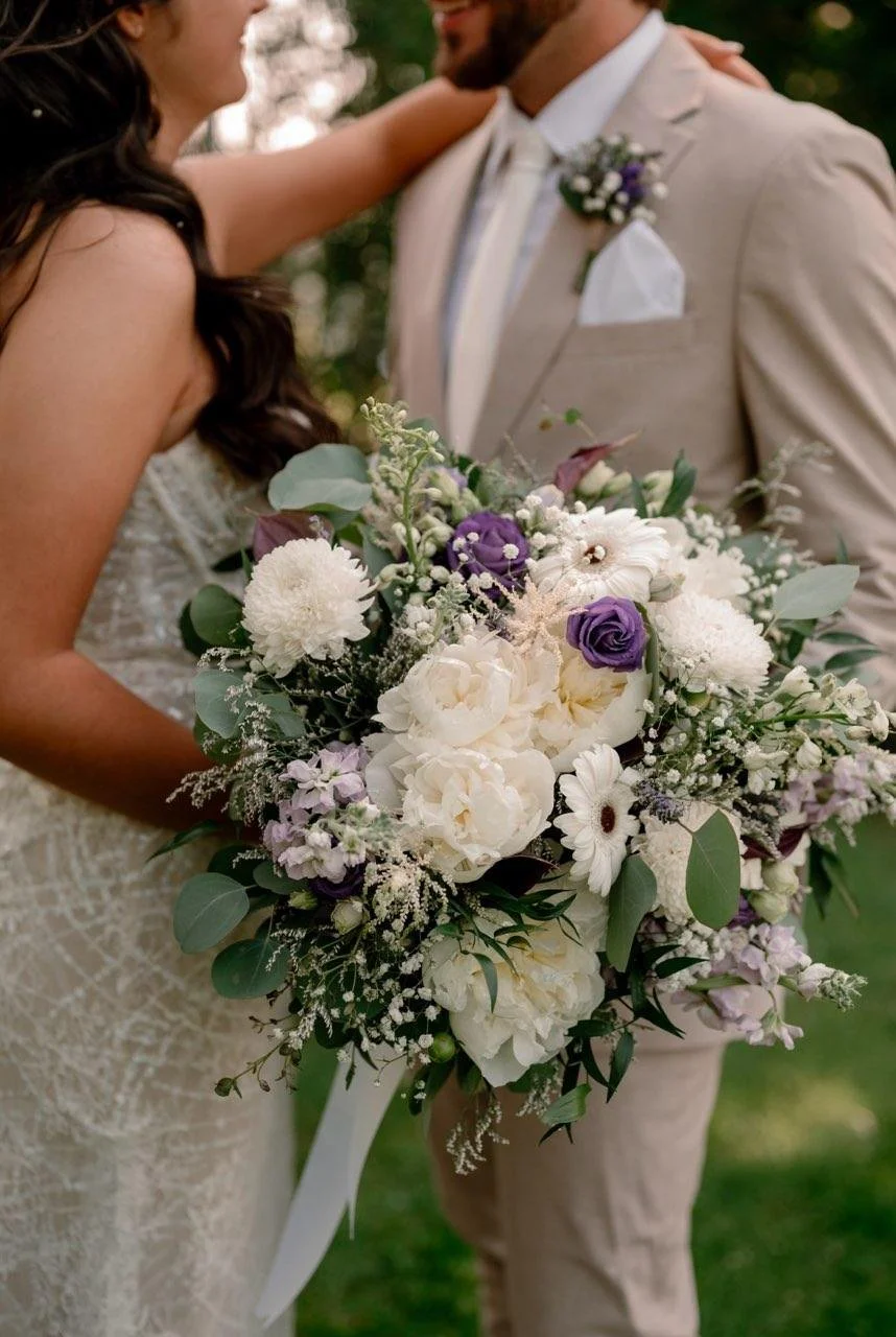 Close-up of a bride and groom holding a large bouquet of white, purple, and lavender flowers at their wedding, outdoors in a park or garden.