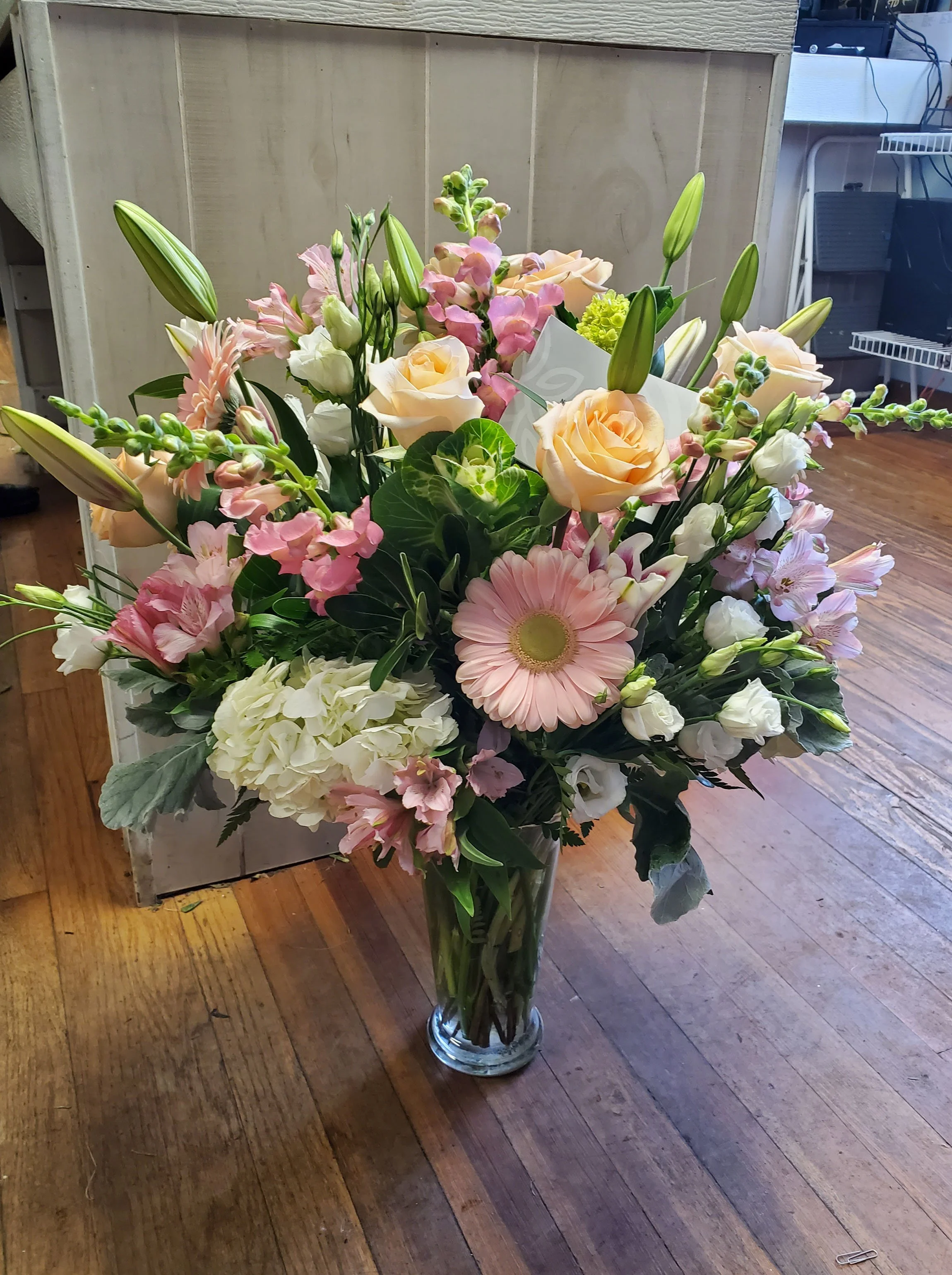 A large bouquet of pink, white, and peach flowers in a tall, clear glass vase on a wooden floor.