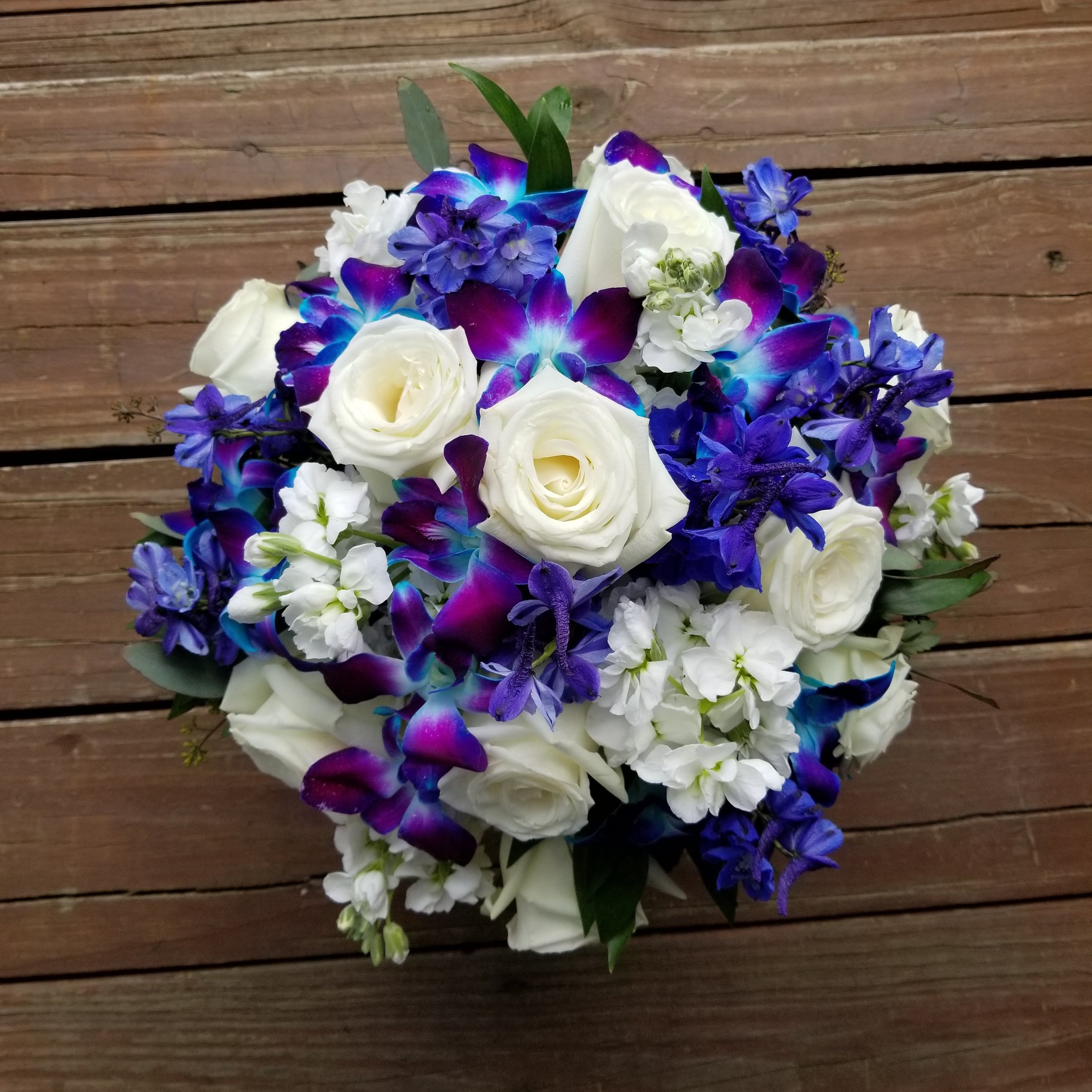 A bouquet of white roses, purple orchids, and other small blue and white flowers on a wooden surface.