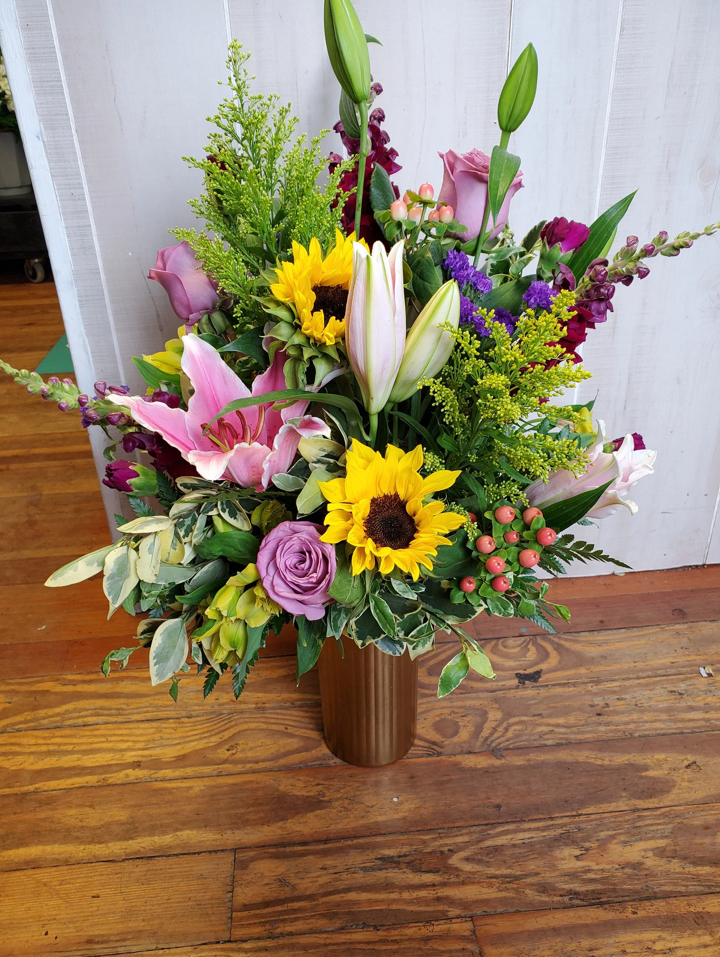 A colorful flower arrangement in a bronze vase, featuring sunflowers, pink lilies, purple roses, lilies, and greenery, placed on a wooden surface.