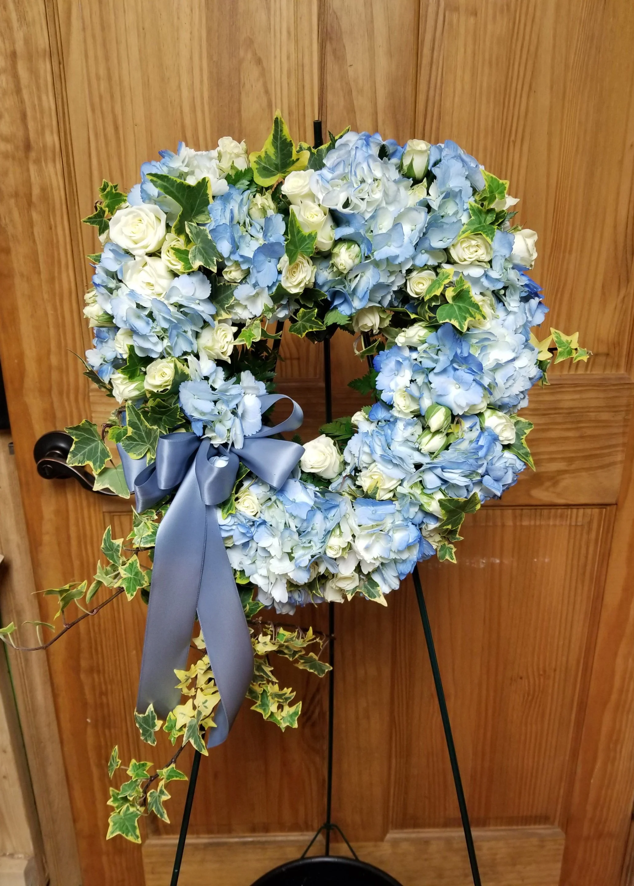 Blue and white floral wreath with a gray ribbon, decorated with ivy, on a stand against a wooden background.