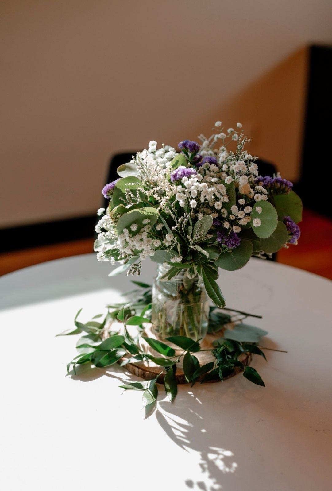 A bouquet of white and purple flowers in a glass jar on a wooden round tray, sitting on a white table with a leafy green vine in front.