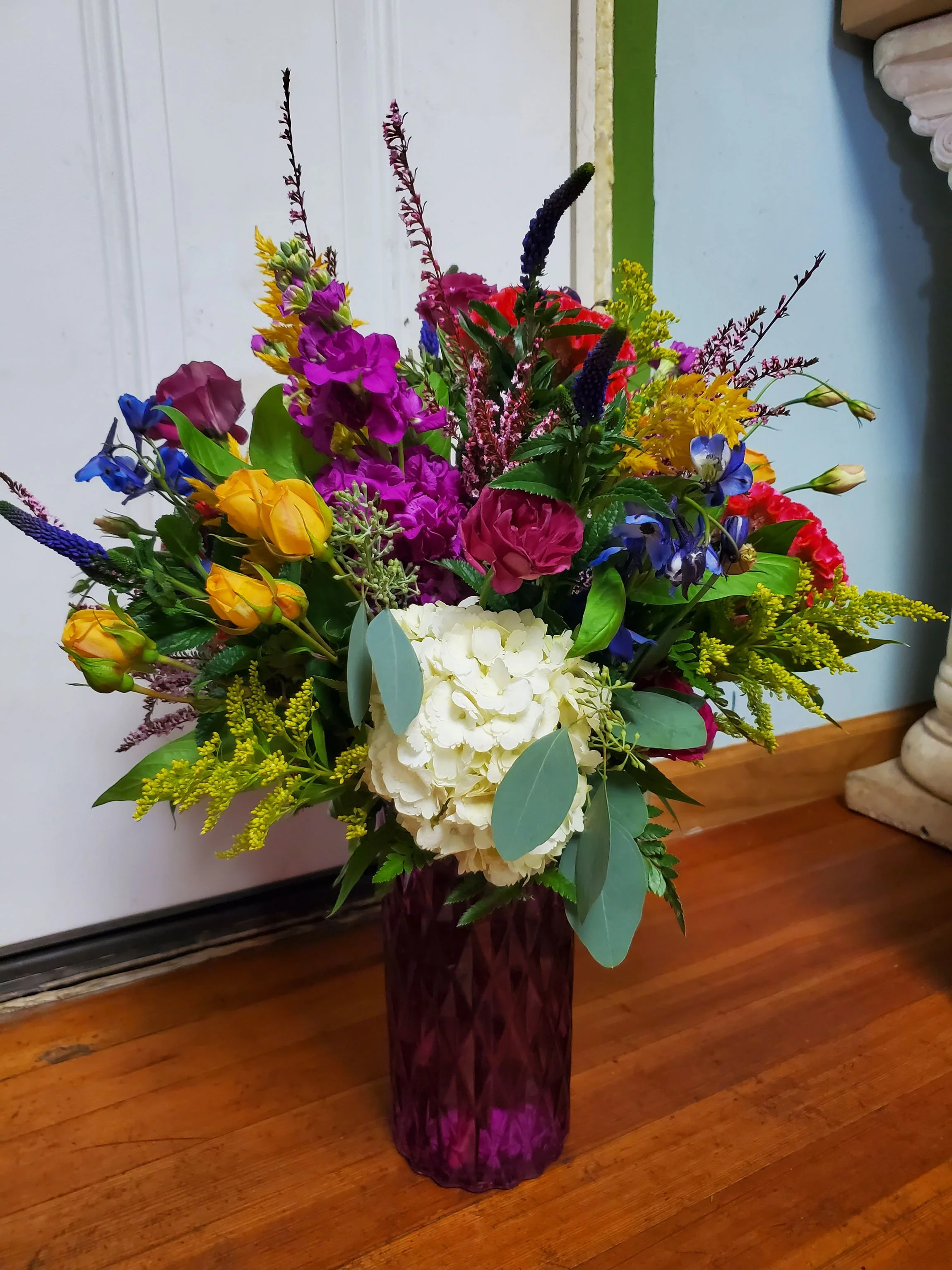 Colorful mixed flower bouquet in a purple textured vase placed on a wooden floor.