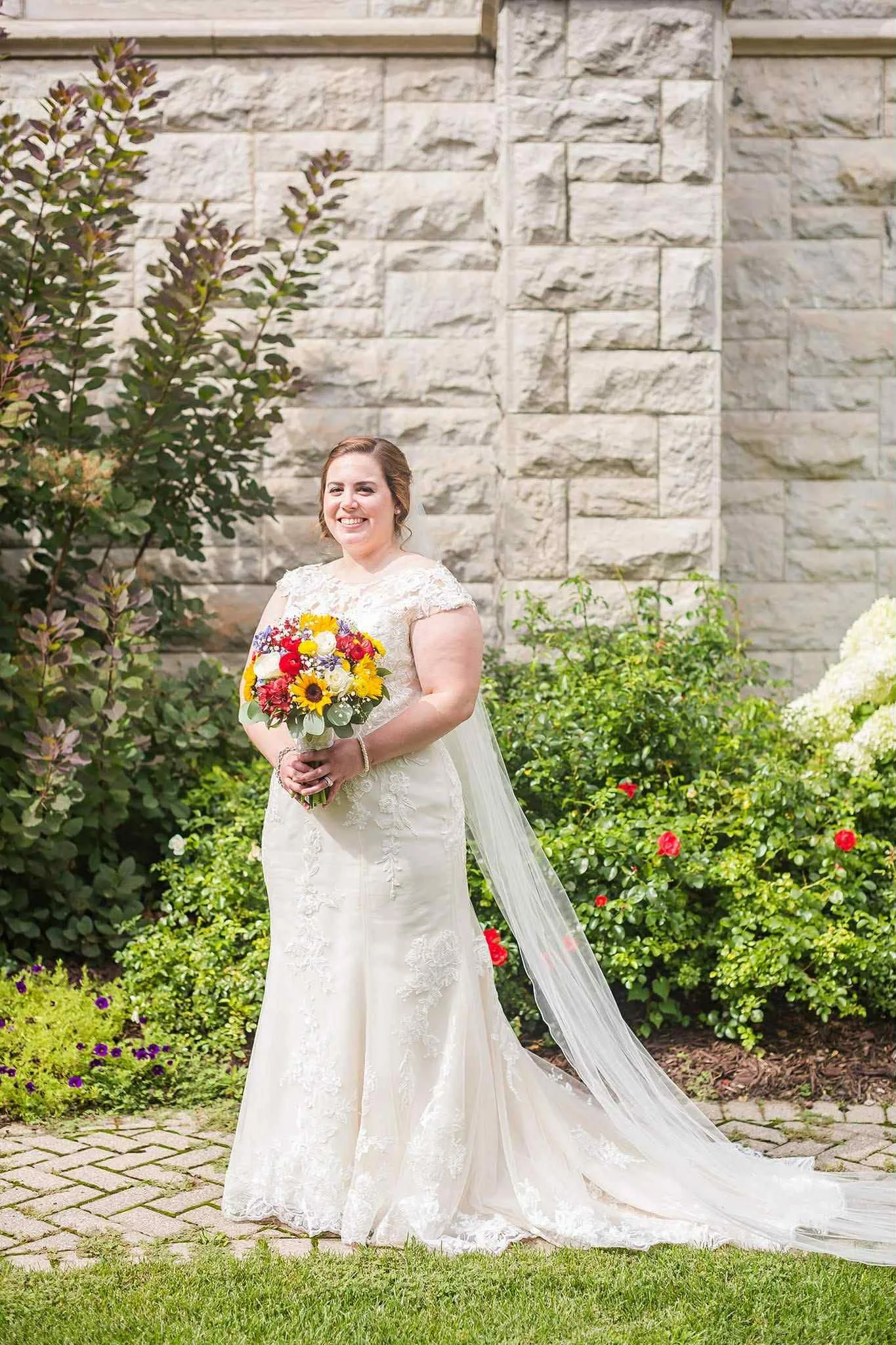 A bride in a white lace wedding gown holding a bouquet of colorful flowers, standing outdoors in front of a stone wall and green bushes.