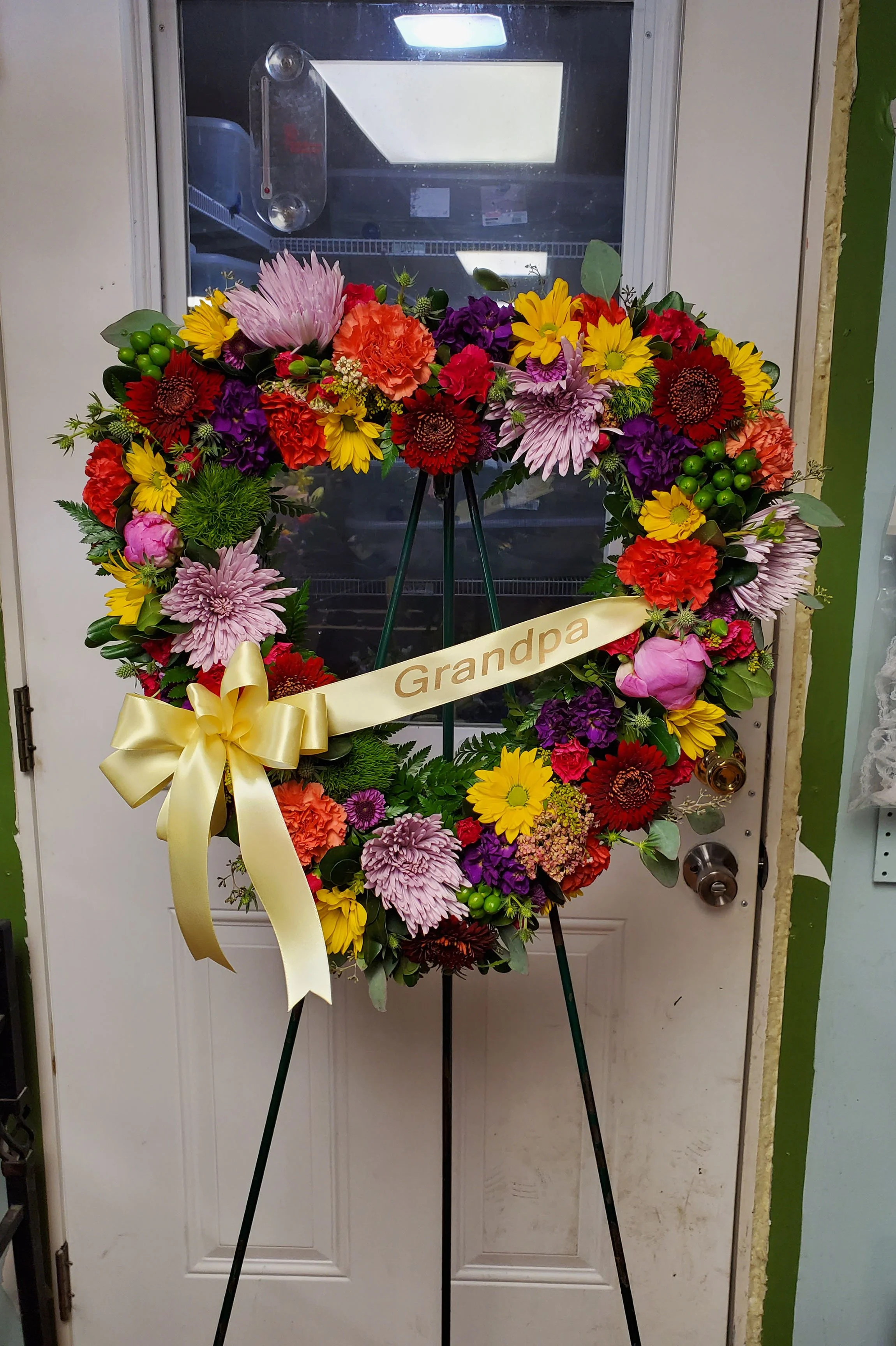Colorful flower wreath on a stand with a yellow ribbon that has the word 'Grandpa' written on it, placed in front of a door.