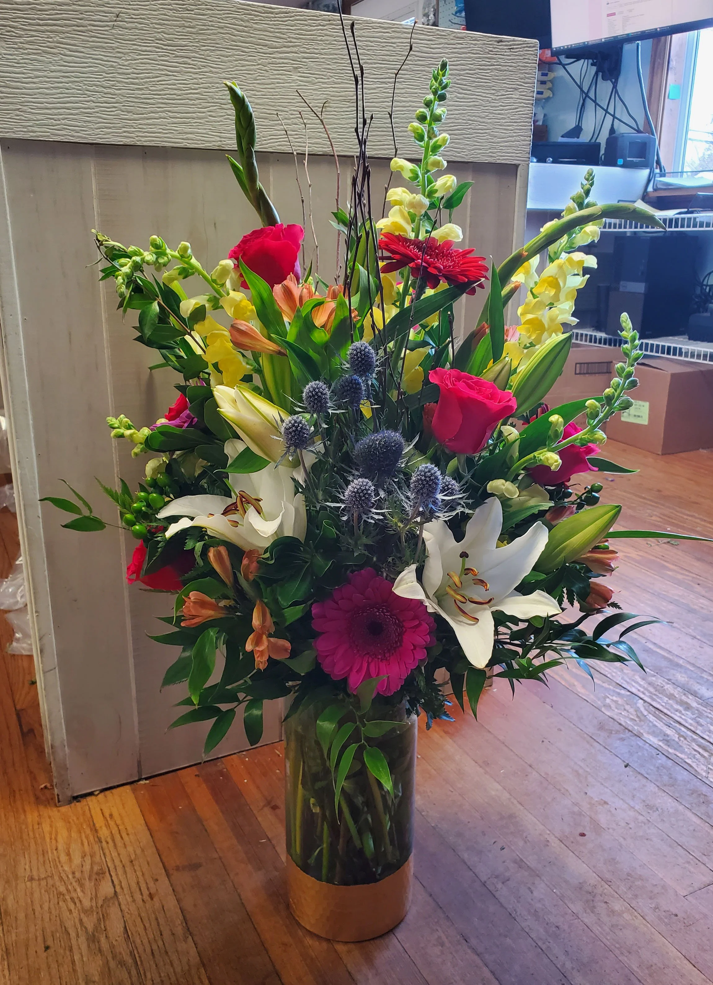 A tall glass vase filled with a colorful arrangement of flowers including red roses, white lilies, pink gerbera daisies, yellow snapdragons, and other green foliage, placed on a wooden floor indoors.