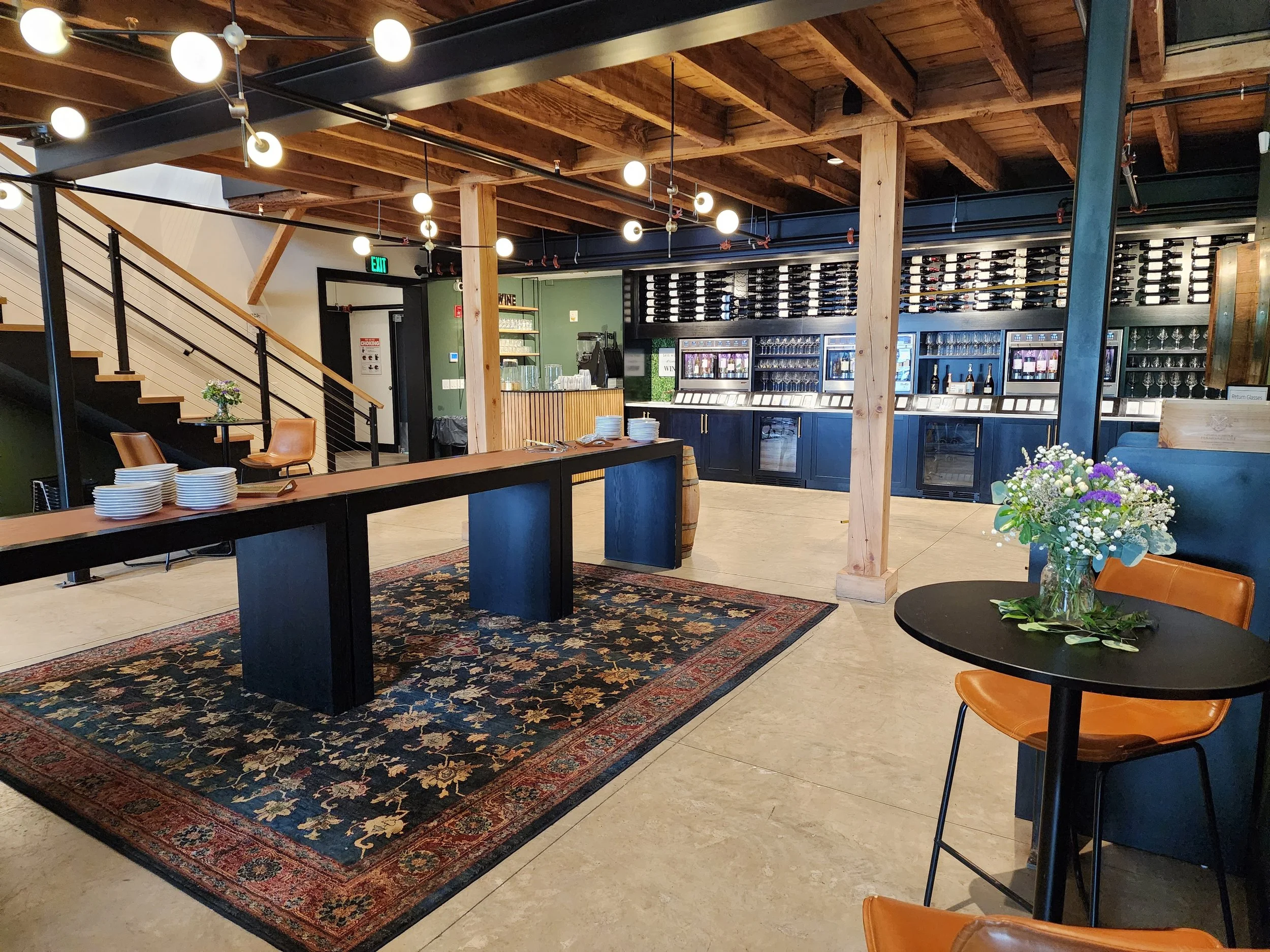 Interior of a modern restaurant or cafe with wooden beams, a patterned rug, and a bar area with wine bottles and glasses. There are tables with chairs, one with a floral arrangement, and a staircase to an upper level.