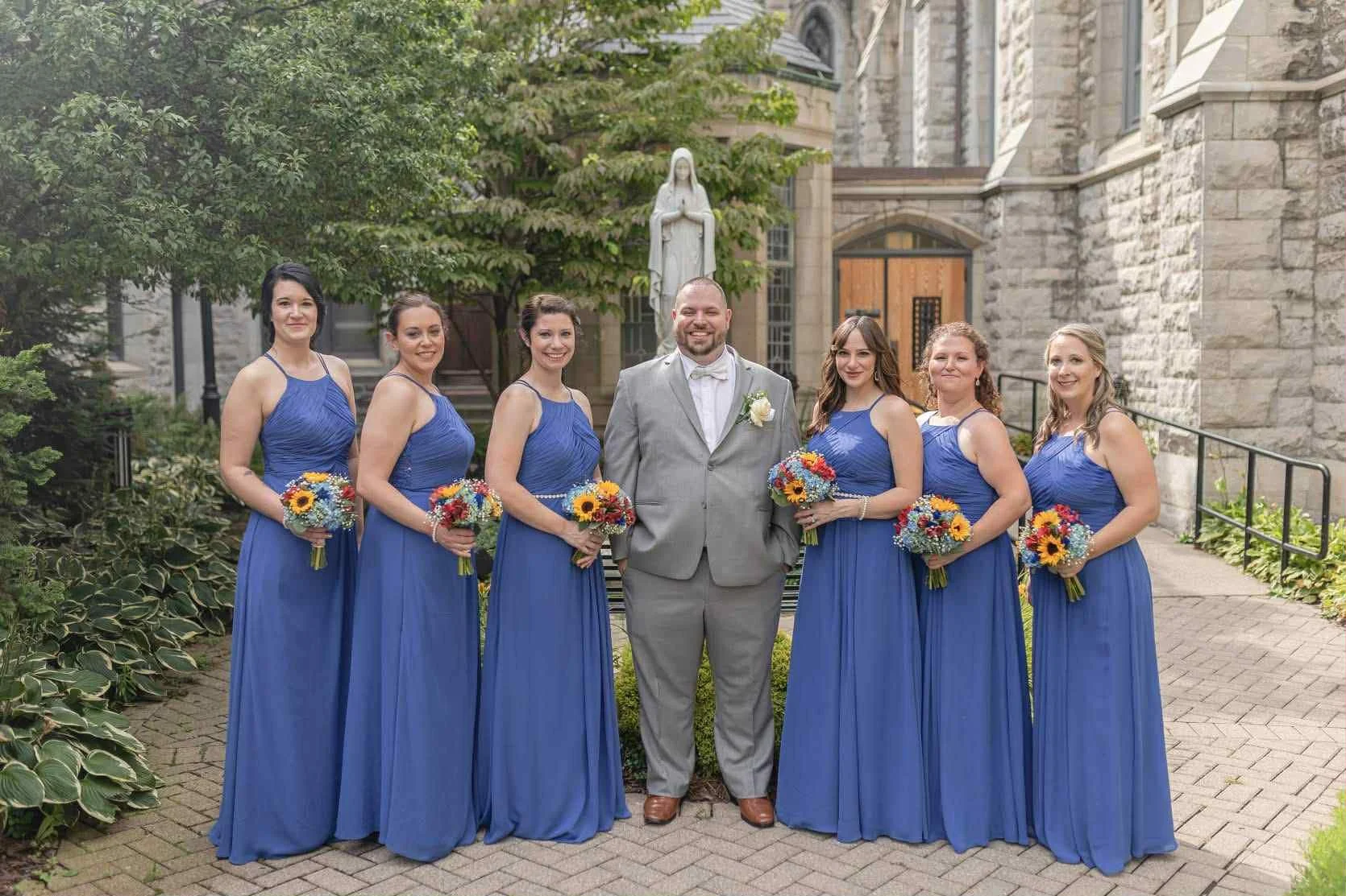 A wedding party outdoors in front of a stone building and statue. Seven women in matching blue dresses and one man in a light gray suit and bow tie are standing in a line, holding bouquets of colorful flowers. The group is smiling, with the man in th