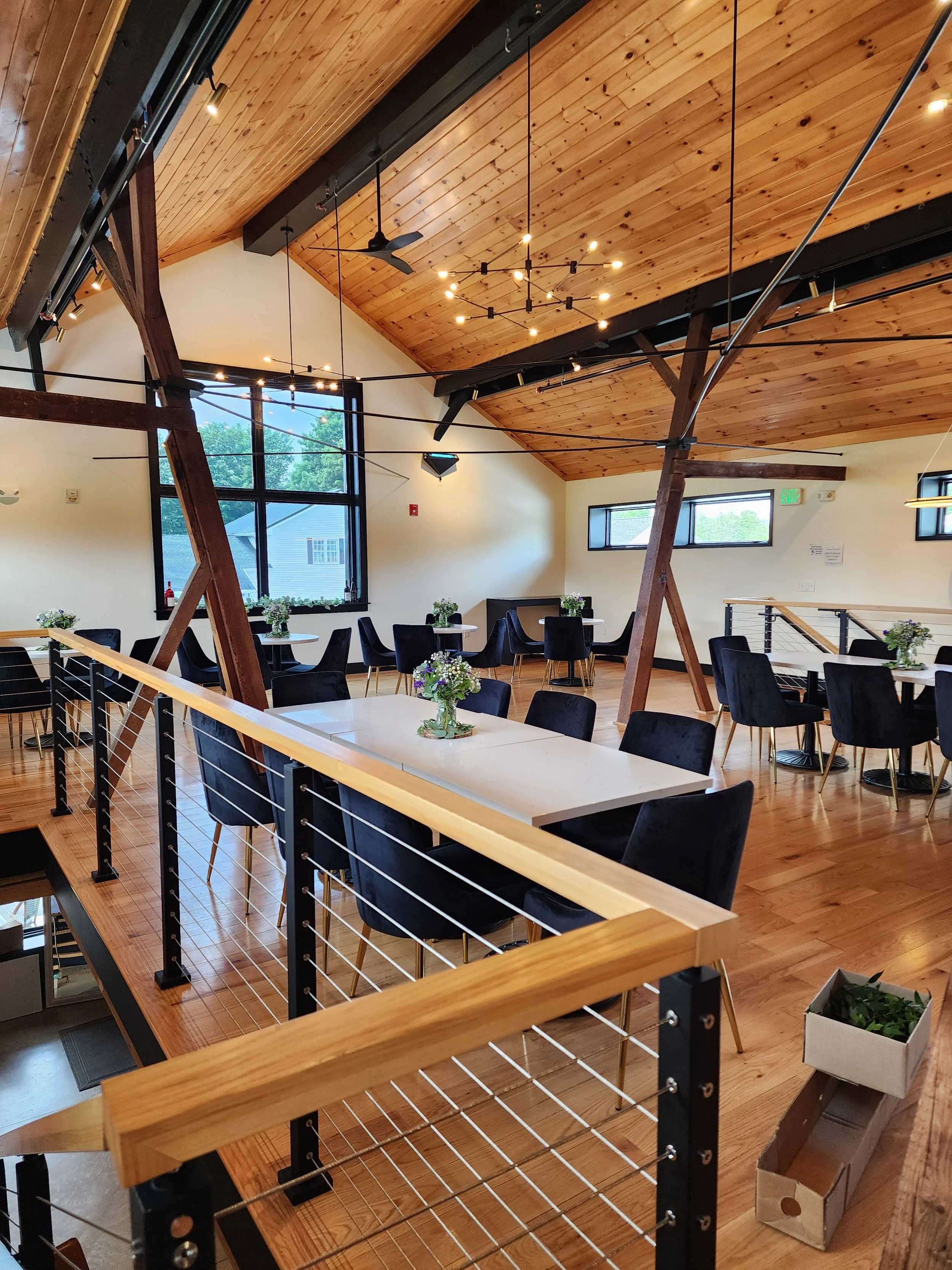 Interior of a modern dining area with wooden ceiling, black chairs, white tables, and decorative plants, featuring large windows and contemporary lighting.